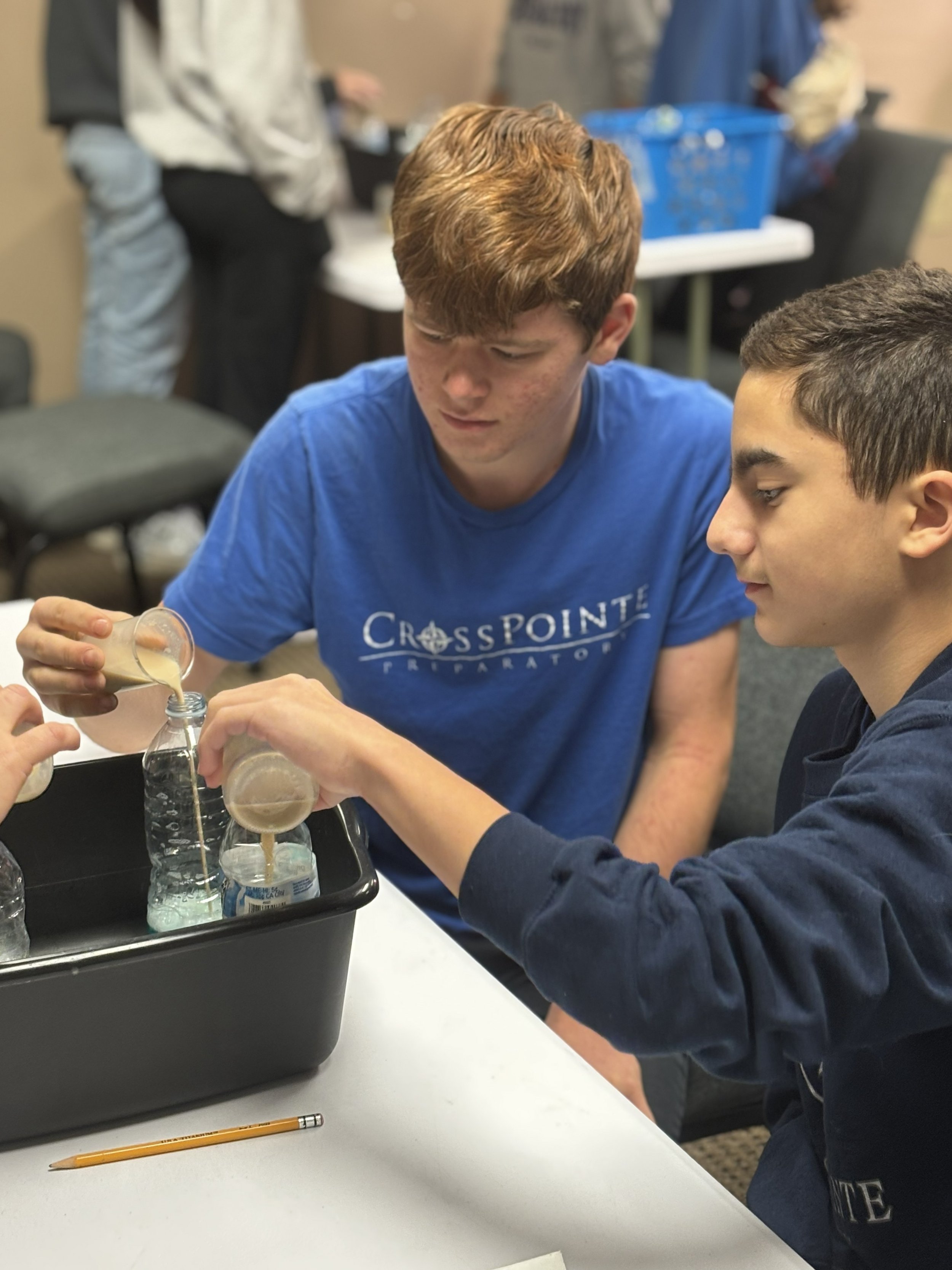 Two boys are demonstrating a science experiment involving pouring liquid from a plastic cup into a ring of water bottles on a table. One boy is wearing a blue CrossPoint shirt, and the other is in a dark-colored hoodie. There is a black tray with water bottles and a yellow pencil on the white table.