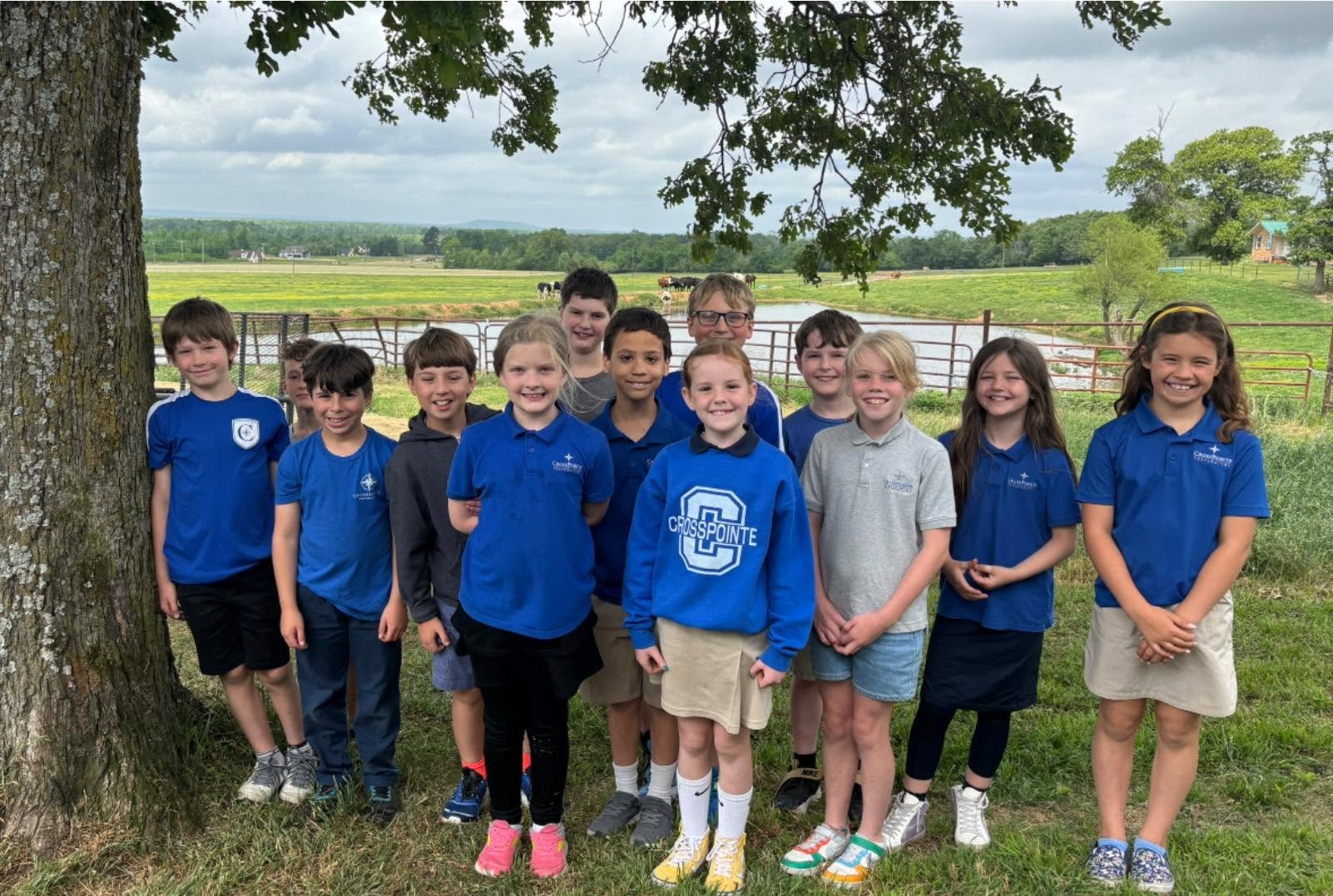 A group of children standing outdoors near a tree, with a farm and pond in the background, smiling and posing for the photo.