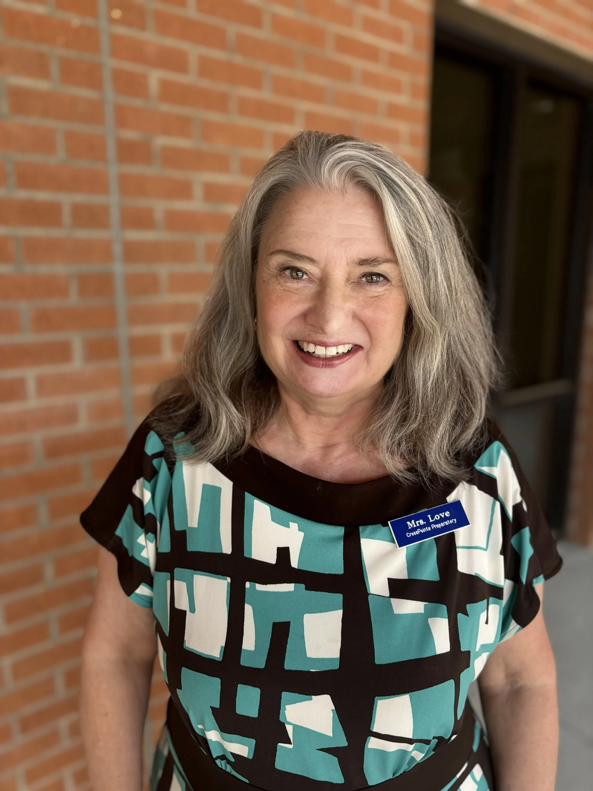 A middle-aged woman with gray hair smiling, wearing a black, white, and teal patterned dress, and a name badge that reads 'Mrs. Love CrossPointe Preparatory'. She is standing outside near a brick wall and a window.