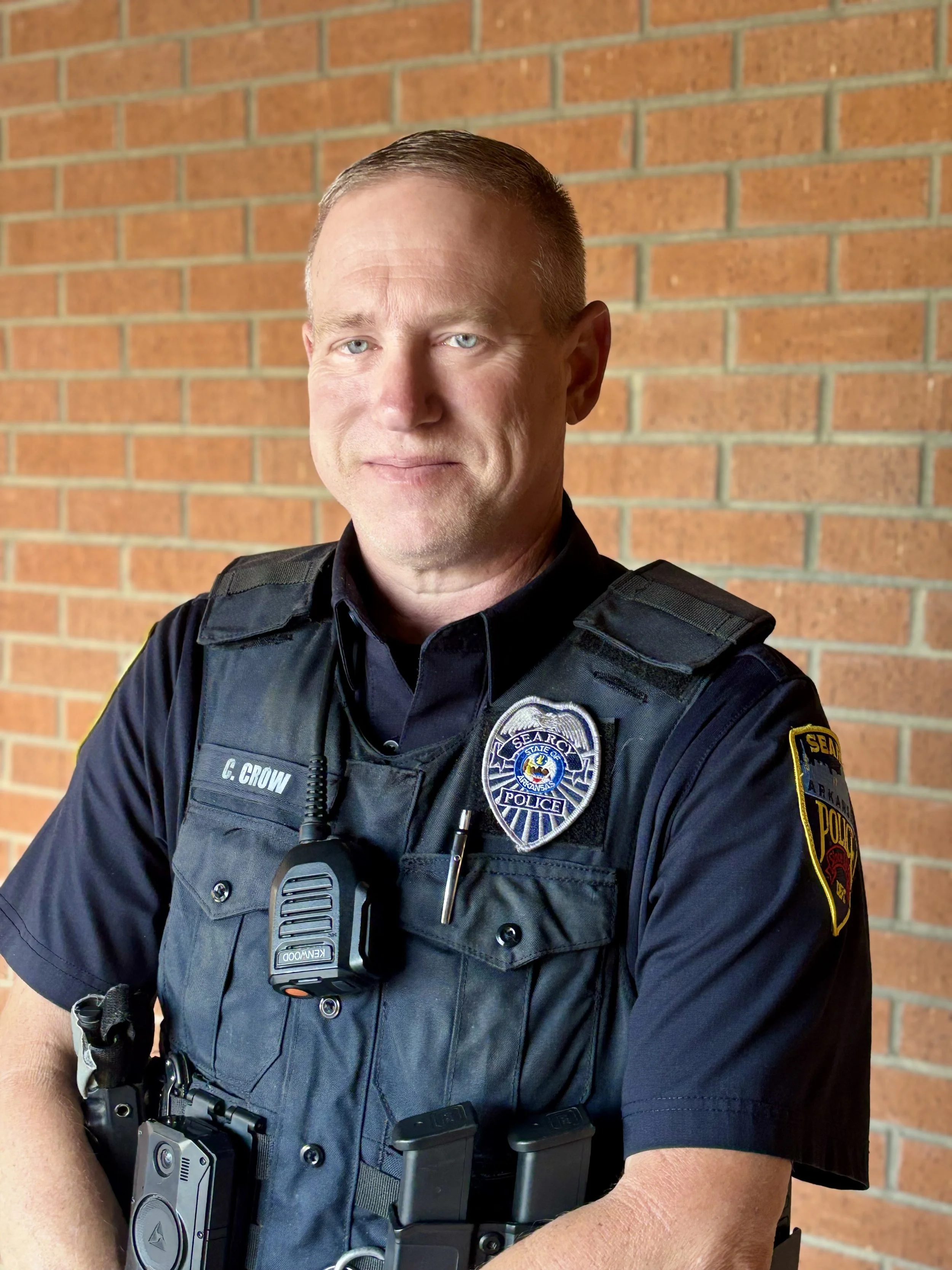 A police officer in uniform standing in front of a brick wall, wearing a body camera, radio, and gear.