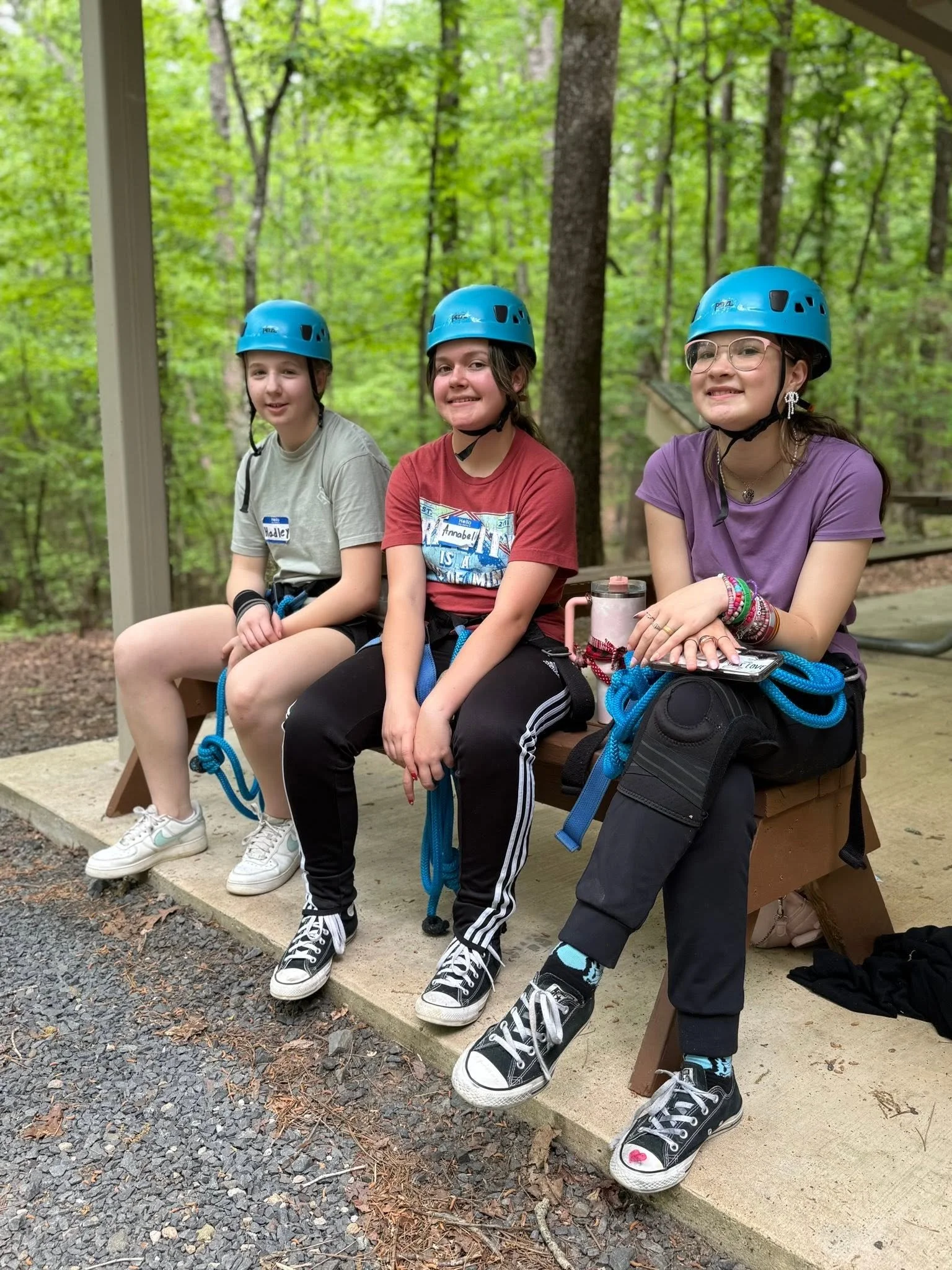 Three young girls sitting on a wooden bench outdoors in a forest, wearing blue helmets and casual clothes.