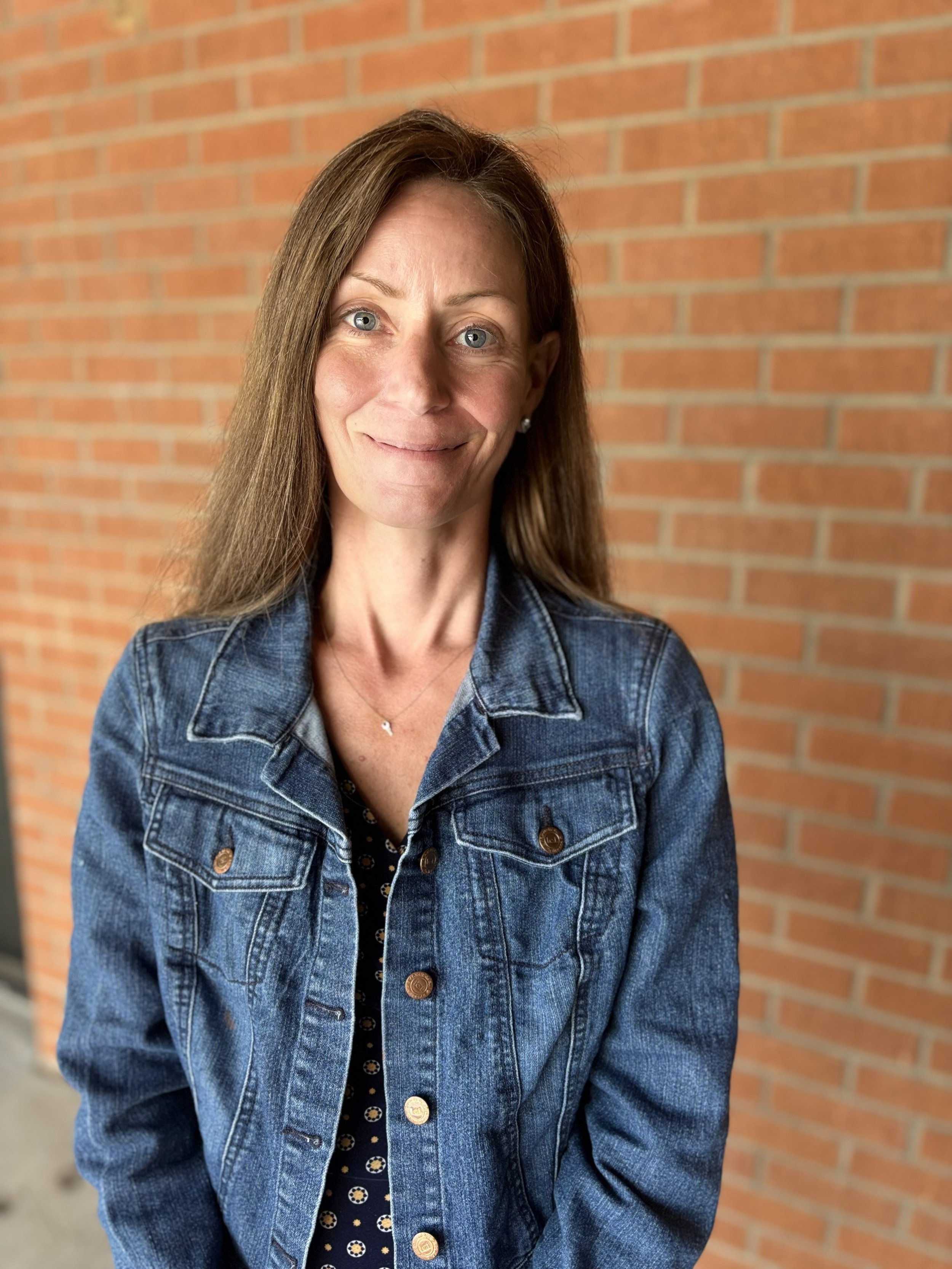 A woman with brown hair and blue eyes wearing a denim jacket and standing against a brick wall.