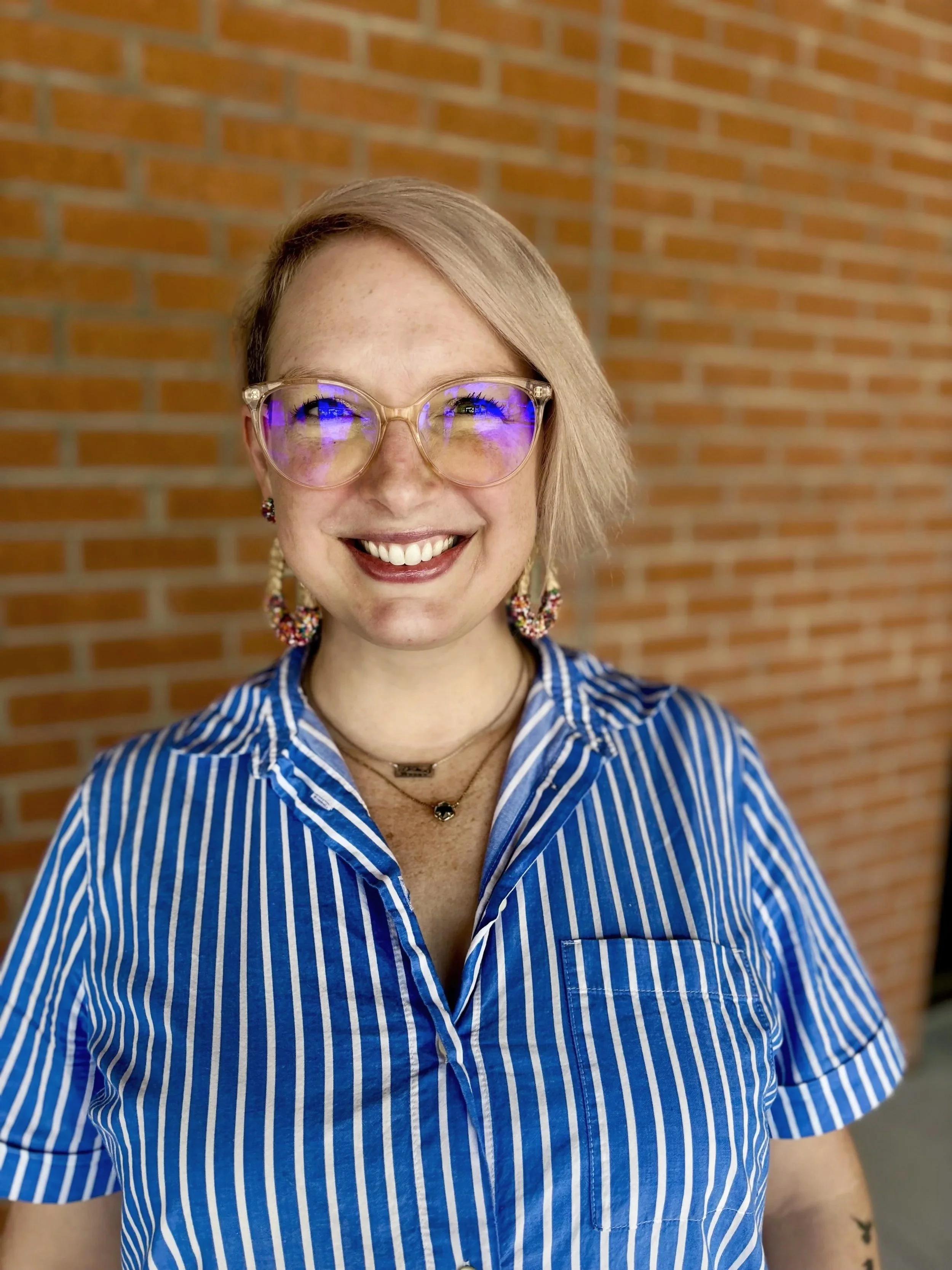 A smiling woman with short blonde hair and glasses with a blue light reflection, wearing a blue and white striped shirt, standing in front of a brick wall.