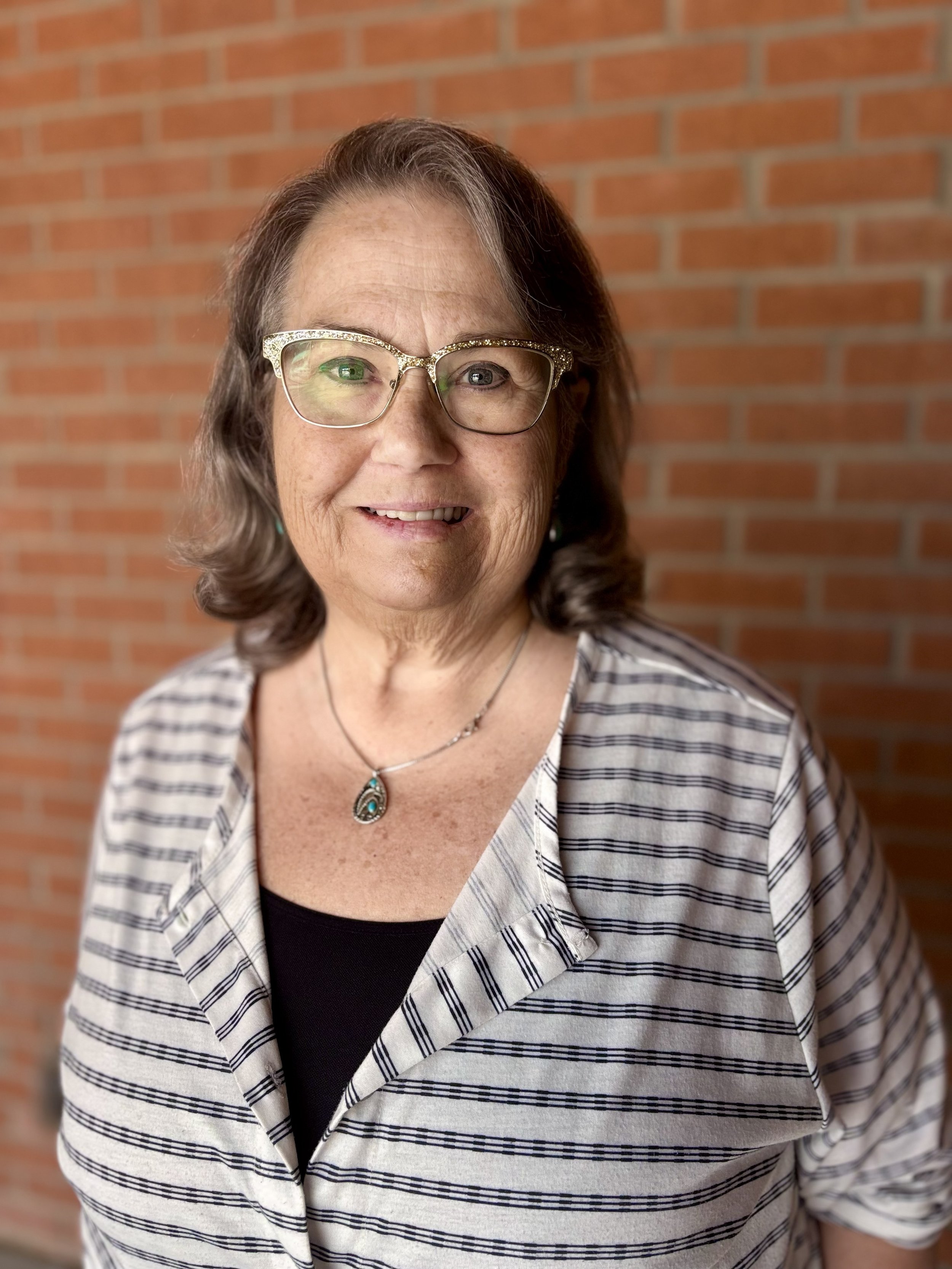 A middle-aged woman with curly brown hair wearing glasses, a striped blazer, and a black top, standing in front of a red brick wall, smiling at the camera.