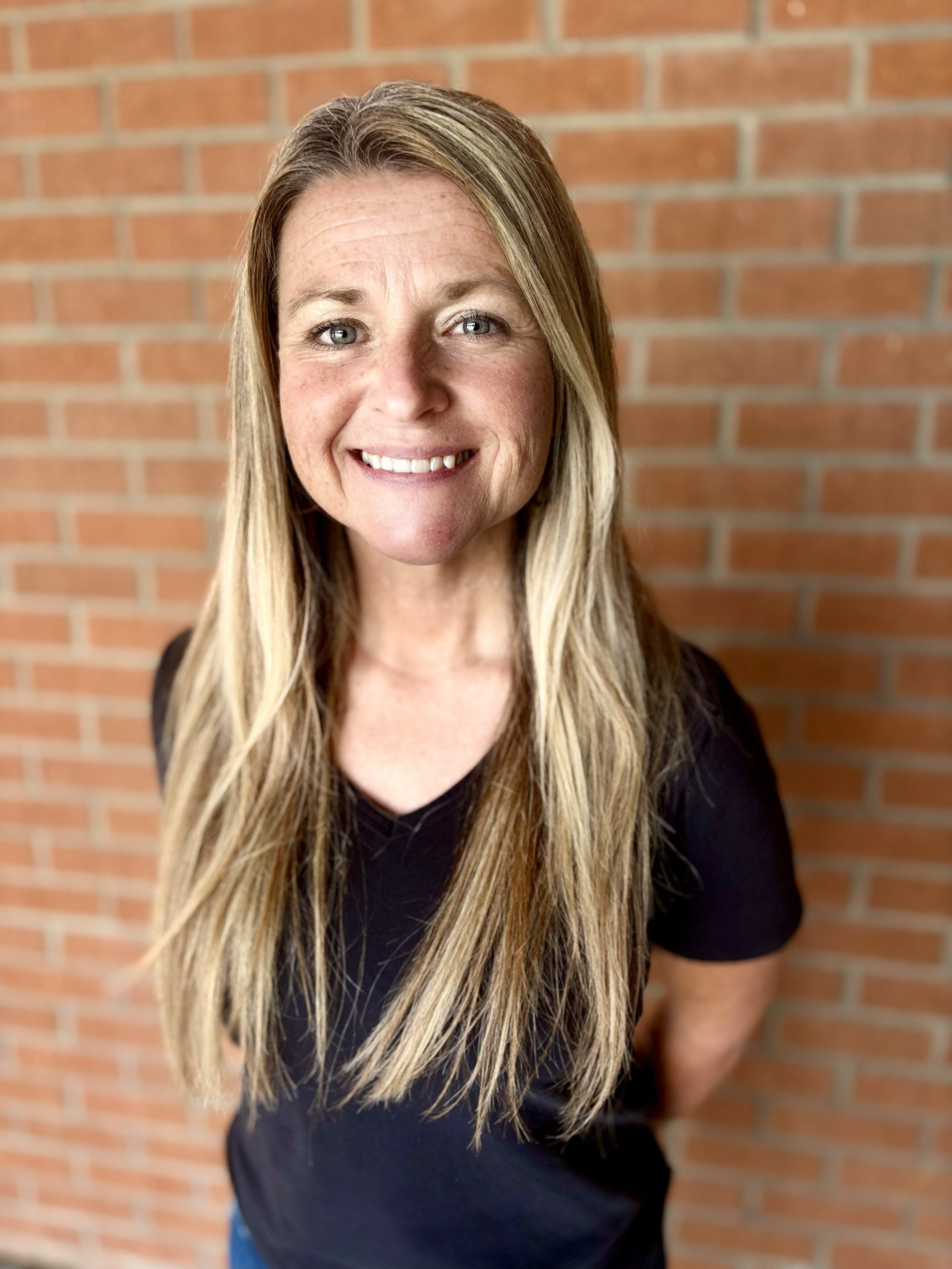 A woman with long blonde hair smiling in front of a brick wall.