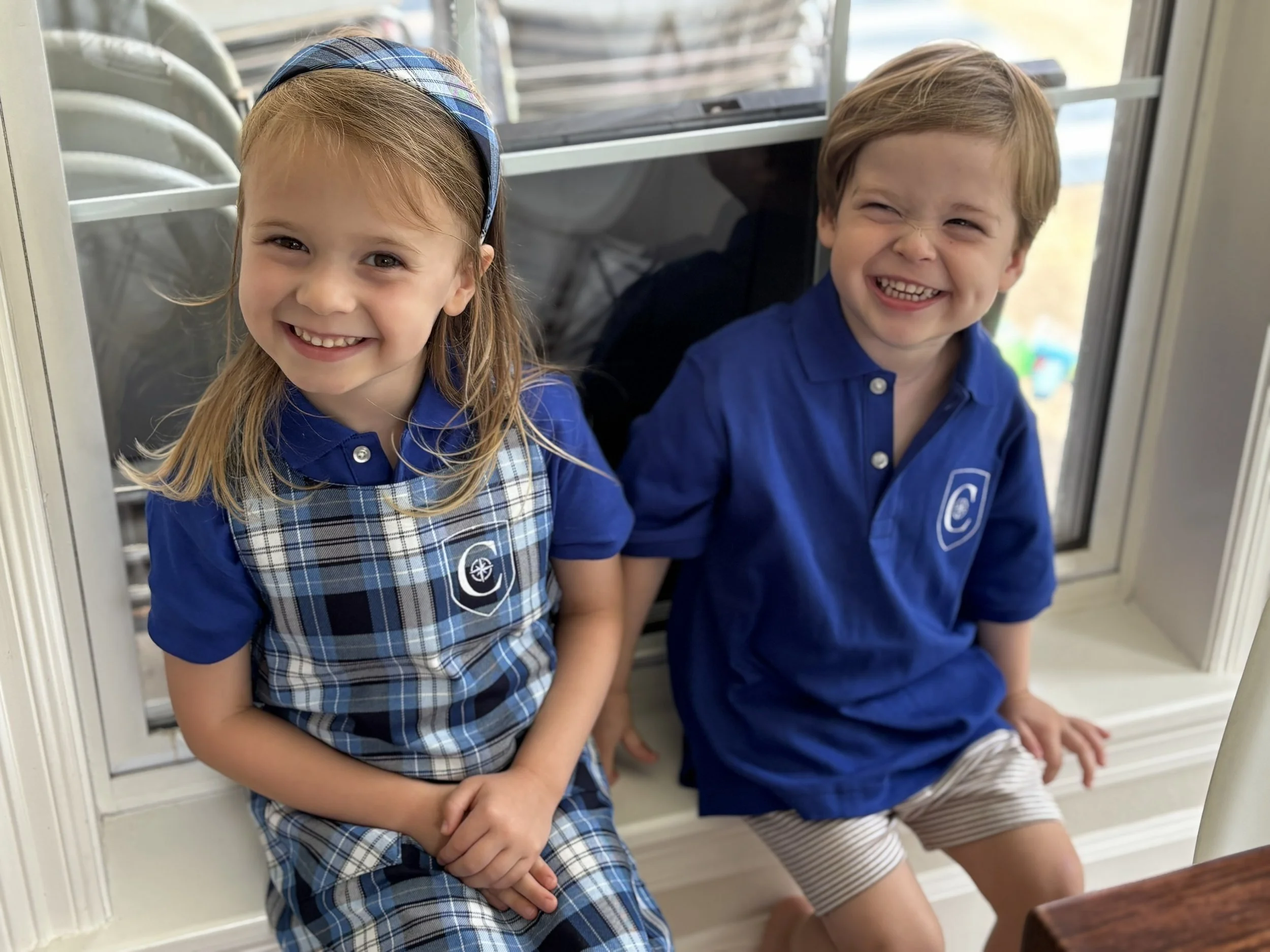 Two young children, a girl and a boy, sitting together near a window. Both are smiling and wearing blue shirts, with the girl also in a plaid dress and headband, and the boy in striped shorts.