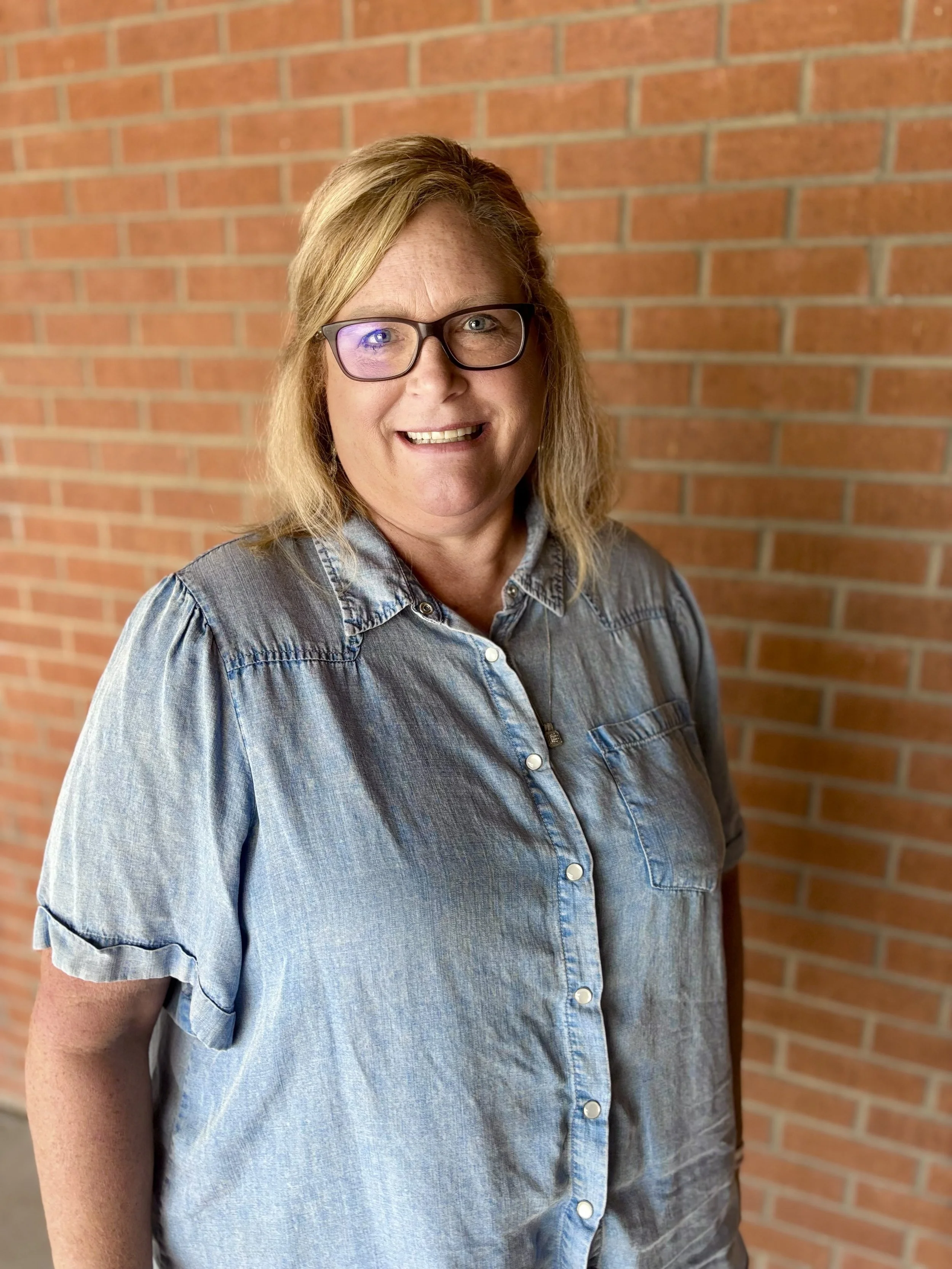 A woman with blonde hair and glasses smiling, wearing a denim shirt against a brick wall background.
