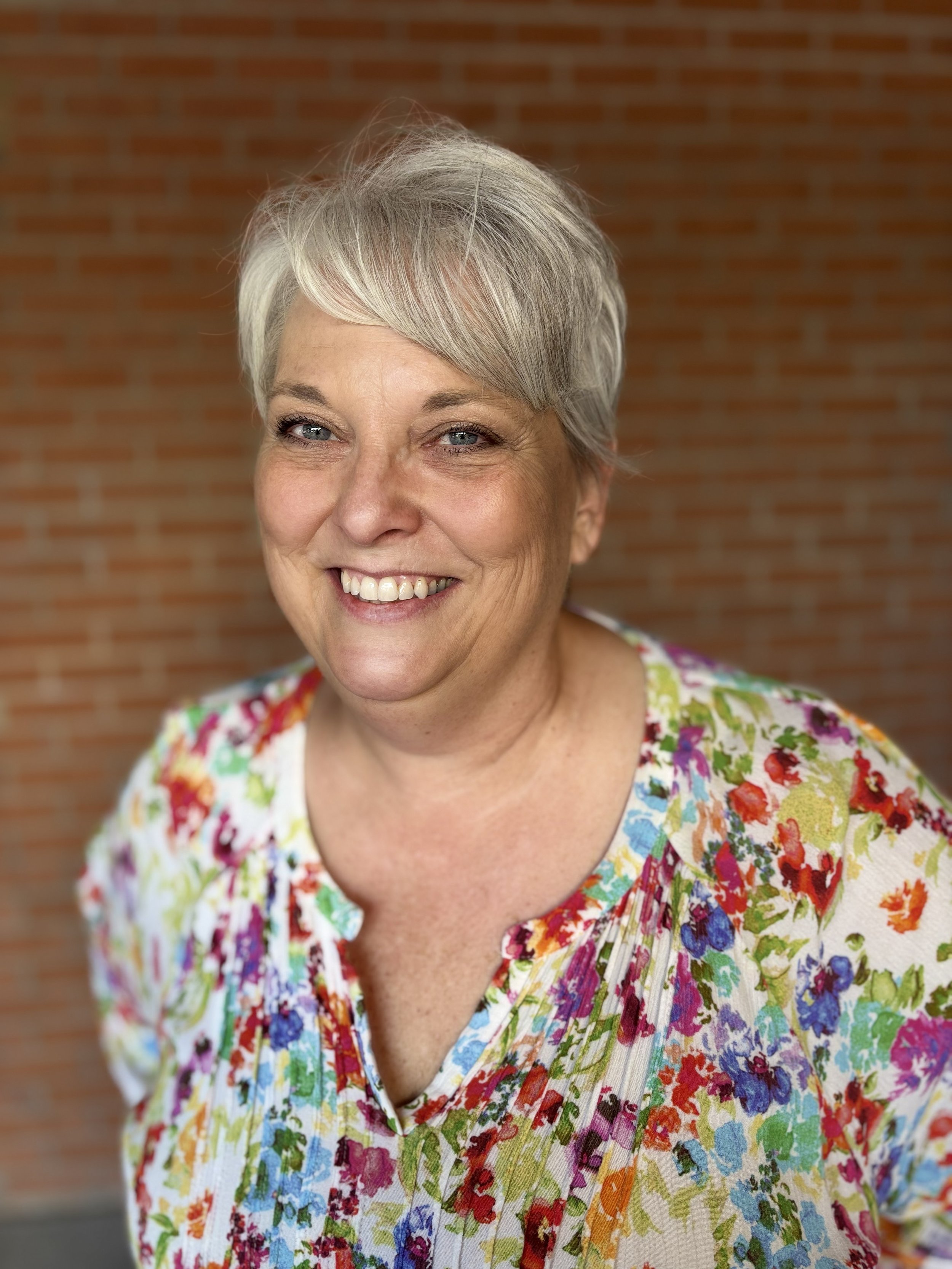 Close-up of a smiling woman with short gray hair, wearing a colorful floral top, in front of a brick wall.