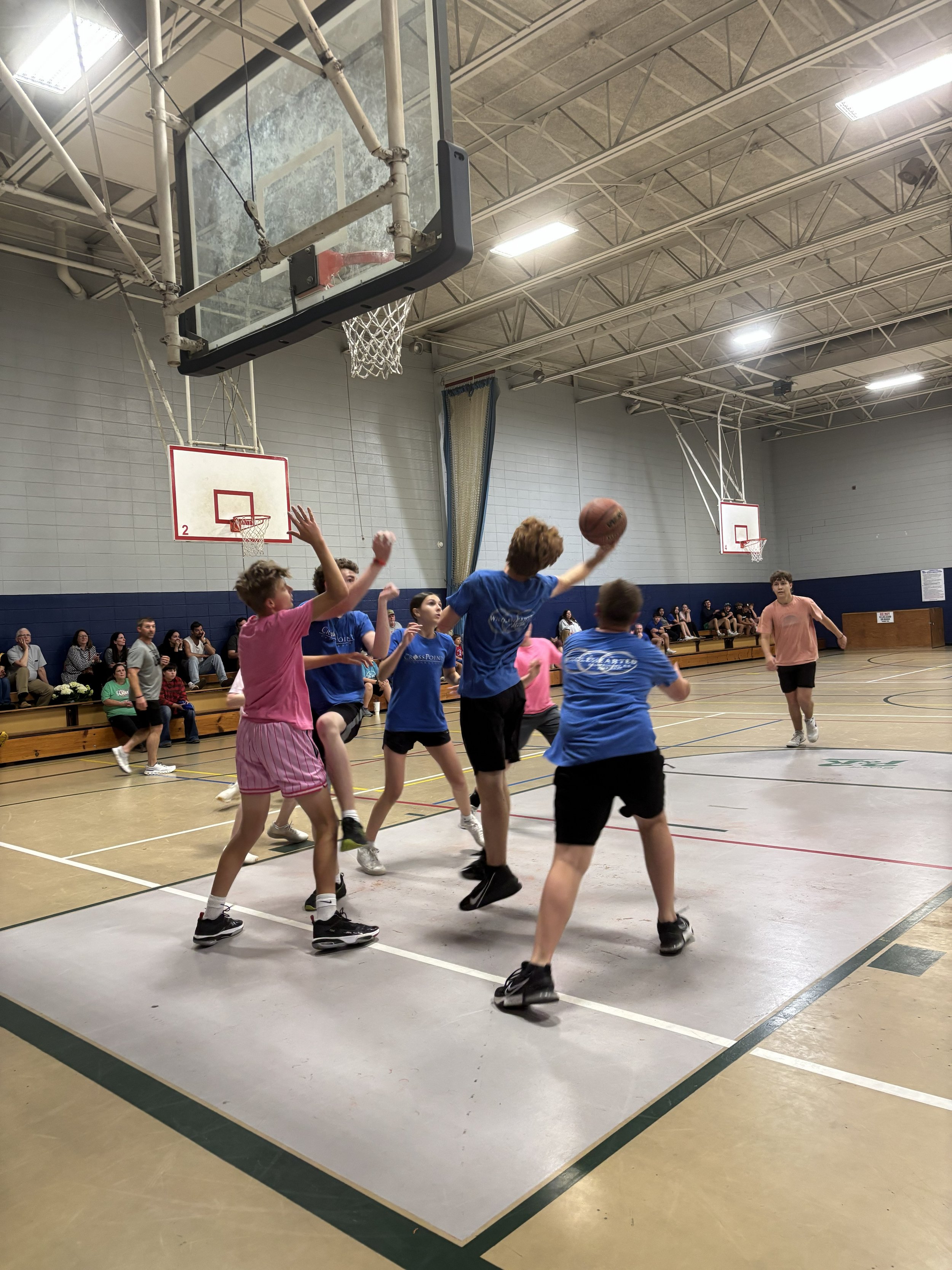 Children playing basketball inside a gym, with spectators seated on benches in the background.