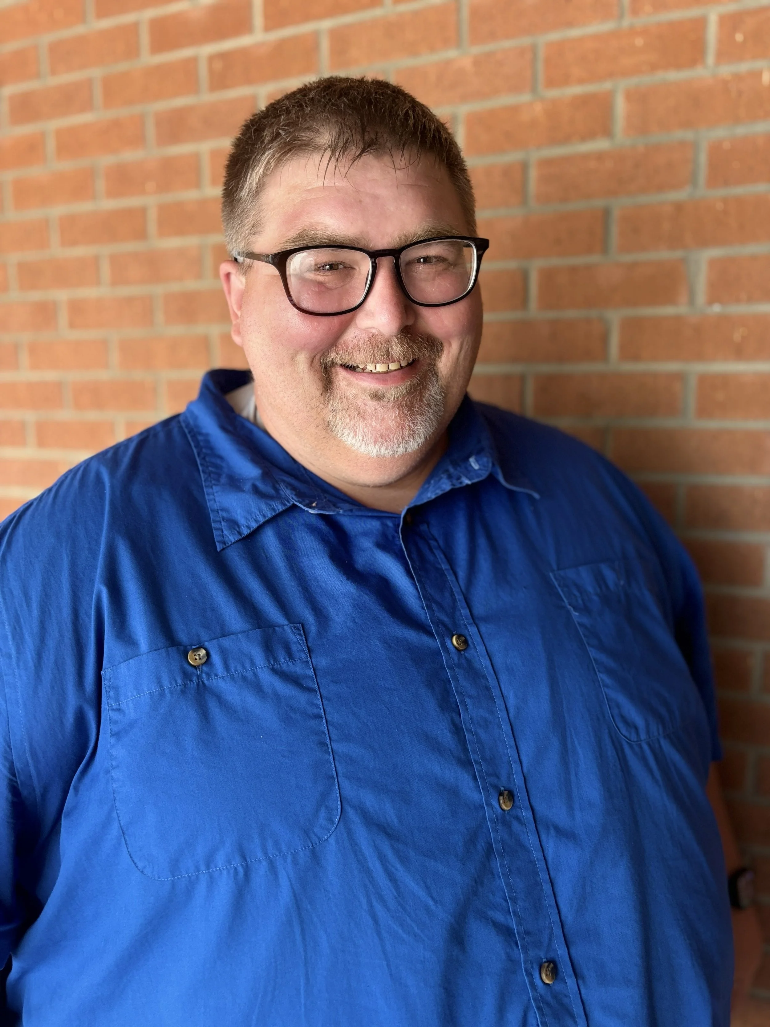 Smiling man with glasses in a blue shirt standing in front of a brick wall.