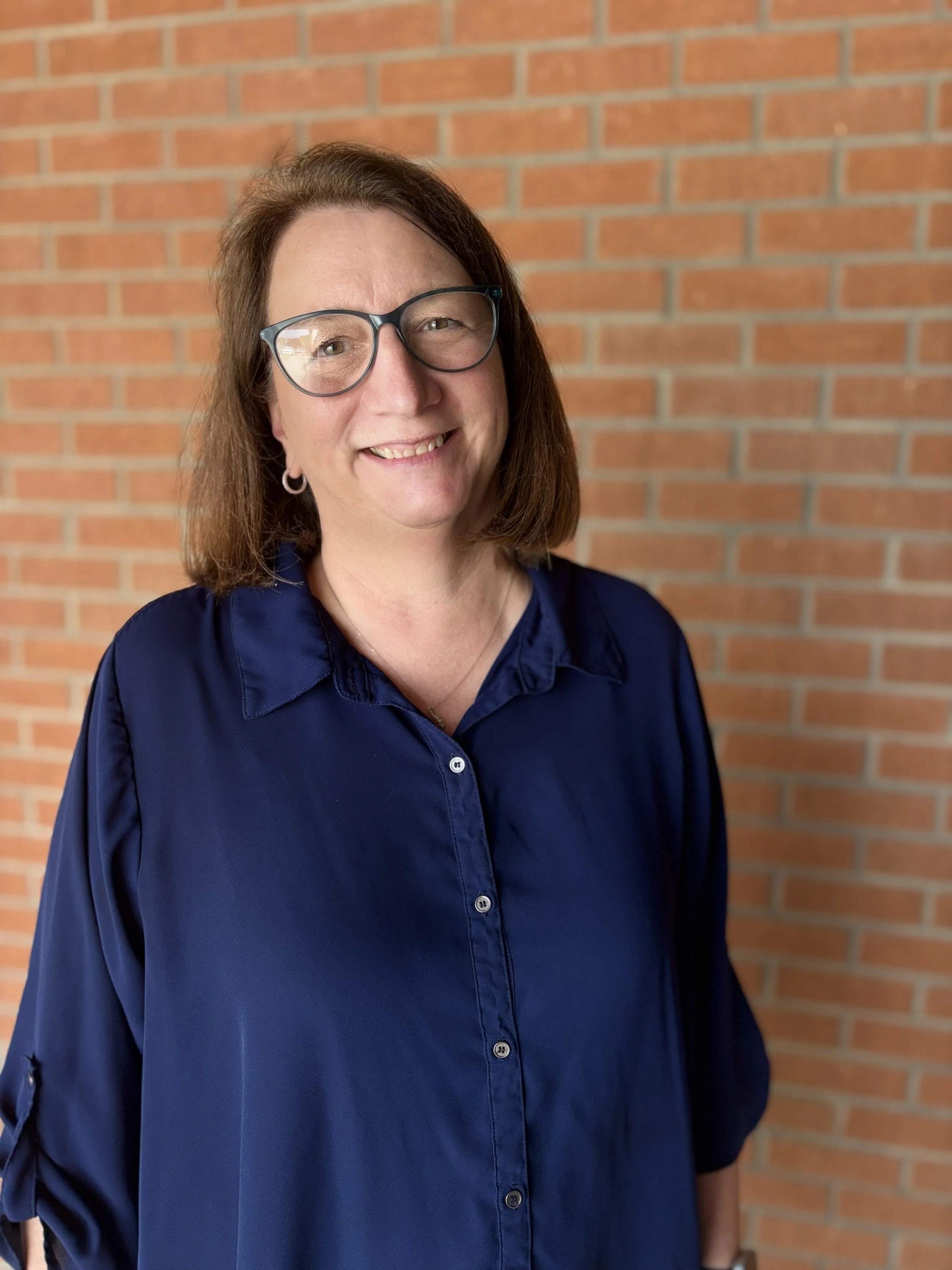 A woman wearing glasses and a navy blue blouse standing in front of a brick wall.