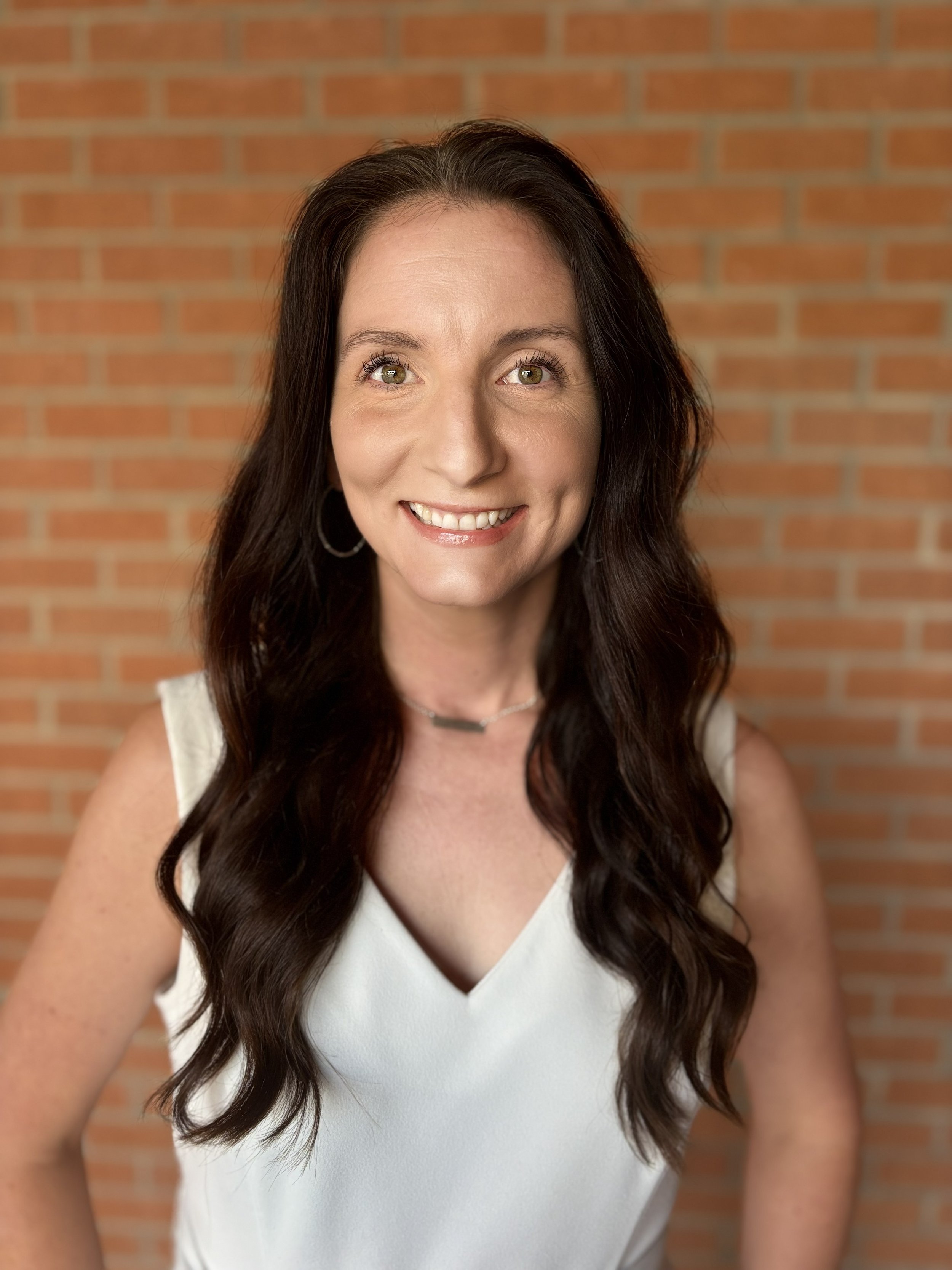 Close-up of a woman with long dark hair, wearing a white top, standing in front of a red brick wall.