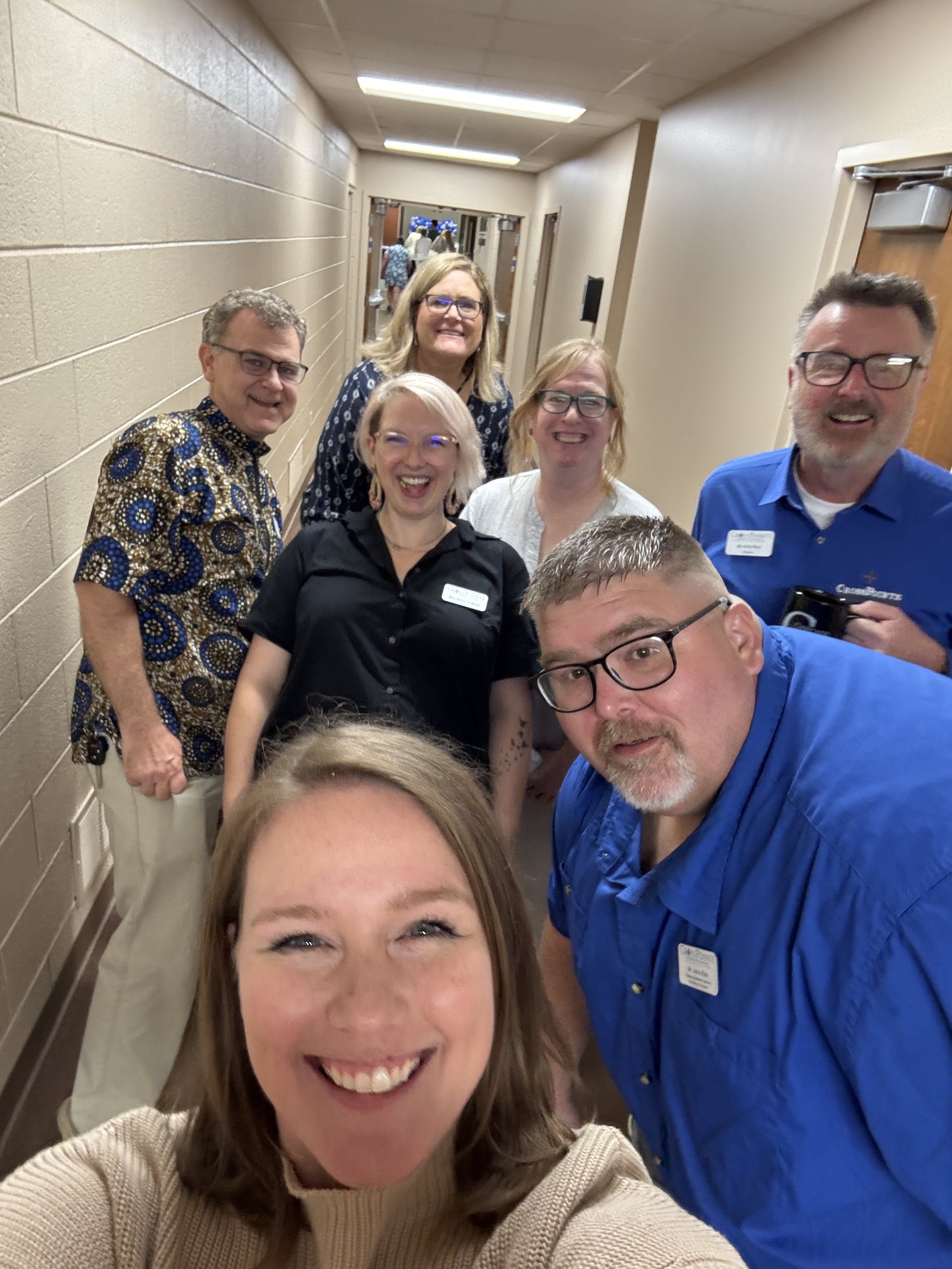 A group of people taking a selfie in a hallway, smiling and wearing casual clothing with name tags, with more people visible in the background.