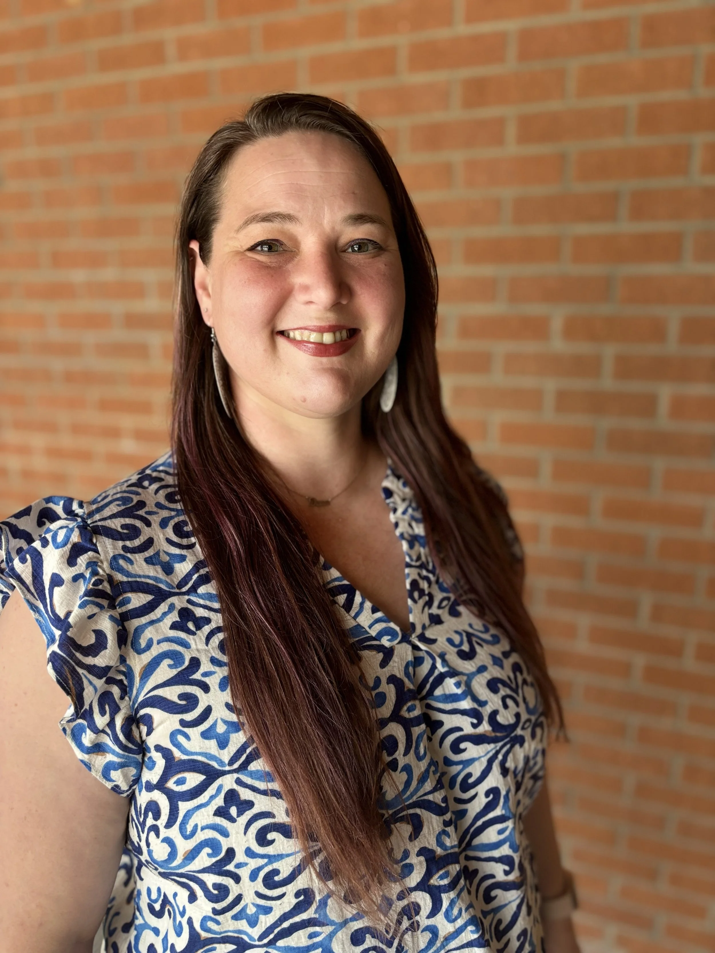 A woman with long brown hair and earrings, smiling, standing in front of a red brick wall, wearing a short-sleeved dress with a blue and beige pattern.
