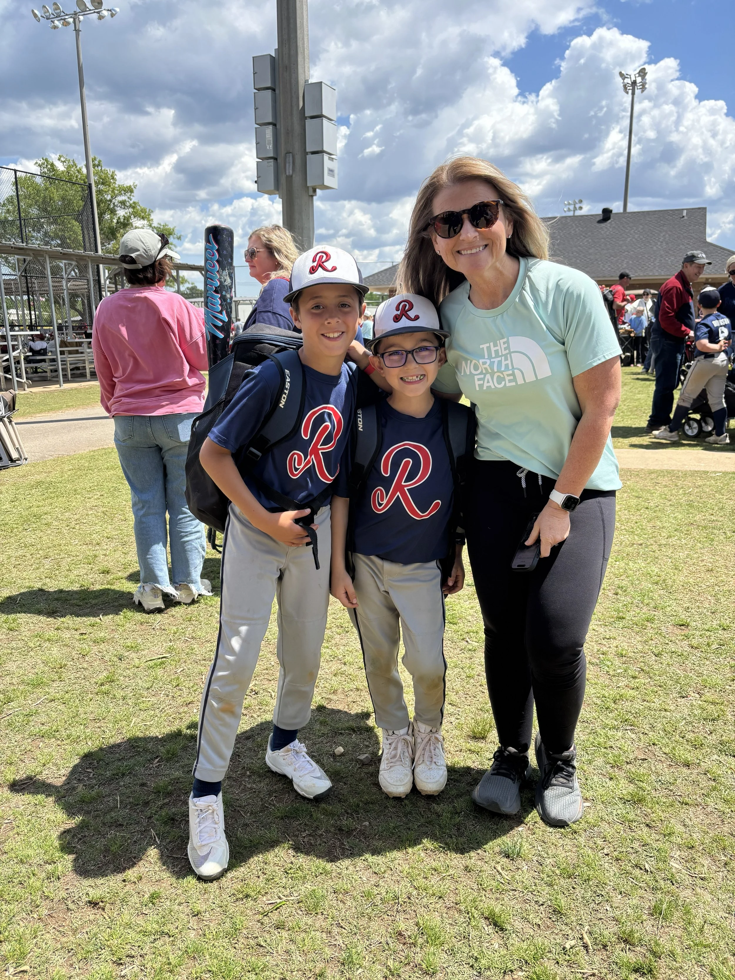 A woman with sunglasses and two boys with matching baseball caps smiling and posing together outdoors at a park or sports field.