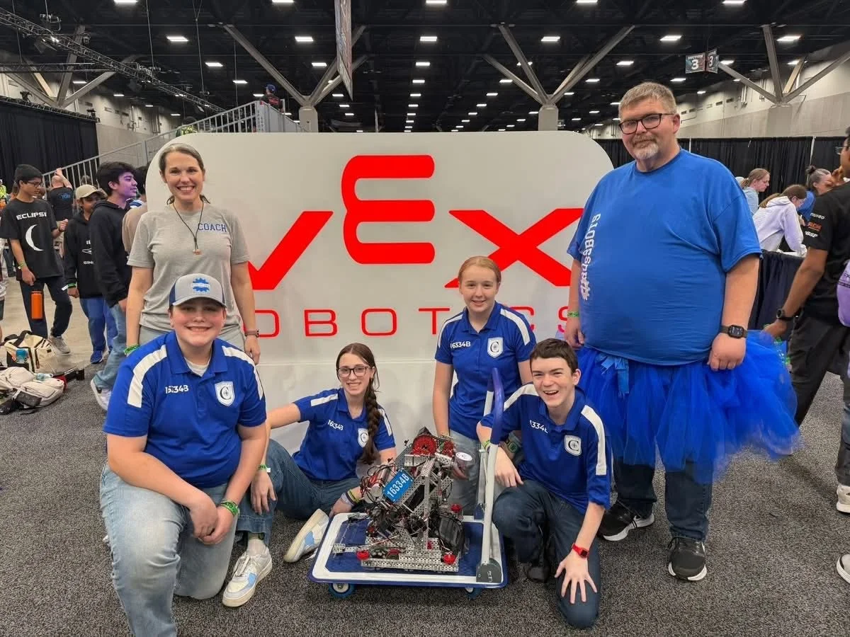 Group of five young robotics team members and their coaches at a robotics competition, with a large sign that reads 'VEX Robotics', and a robot in front of them.