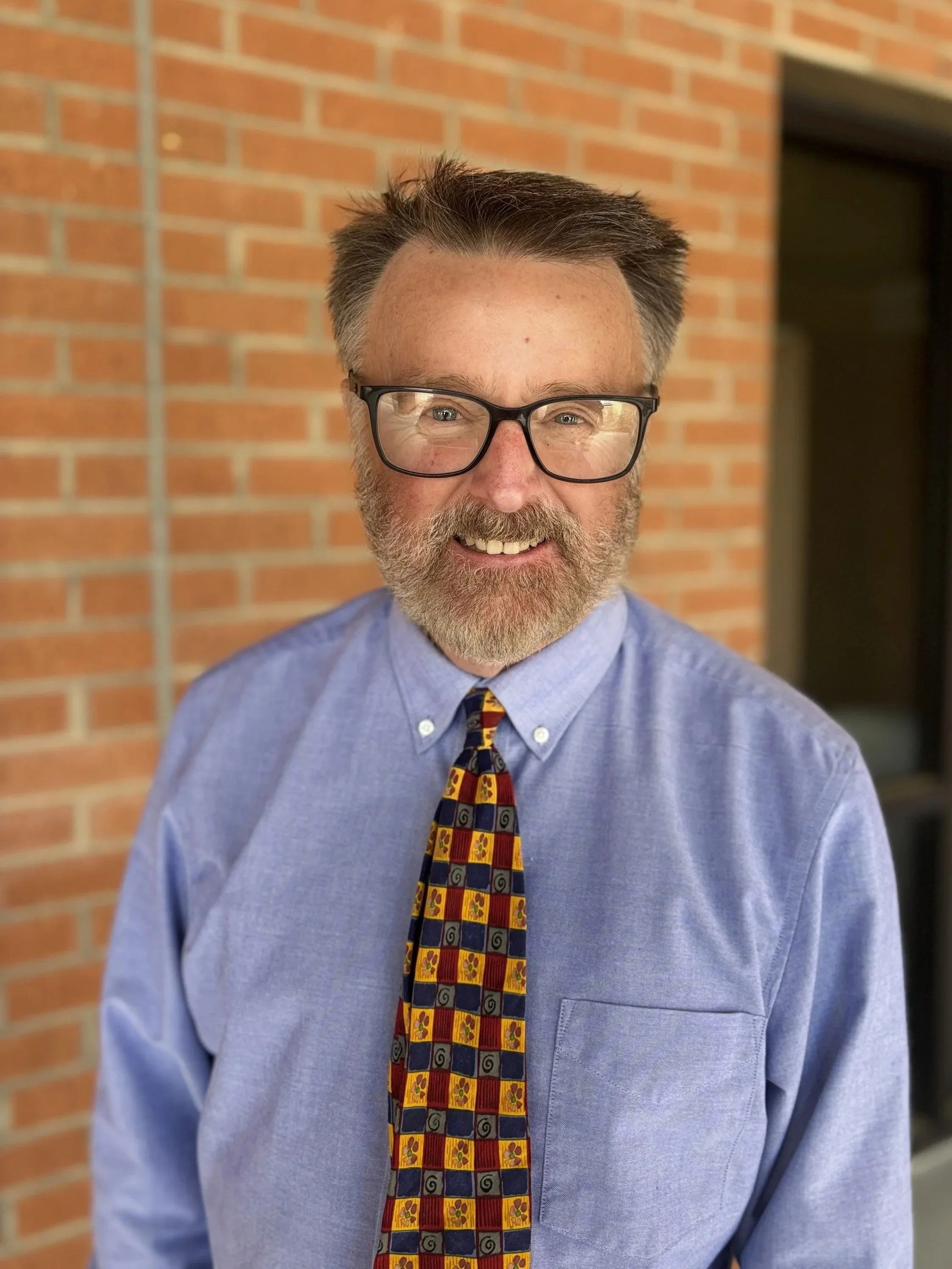A smiling man with glasses and a beard wearing a light blue shirt and a colorful patterned tie, standing in front of a brick wall.