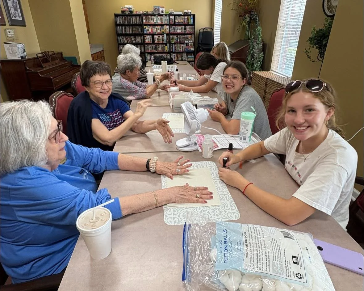 A group of women and young girls sitting around a table in a room, engaged in a nail painting or skincare activity, with smiles and laughter.