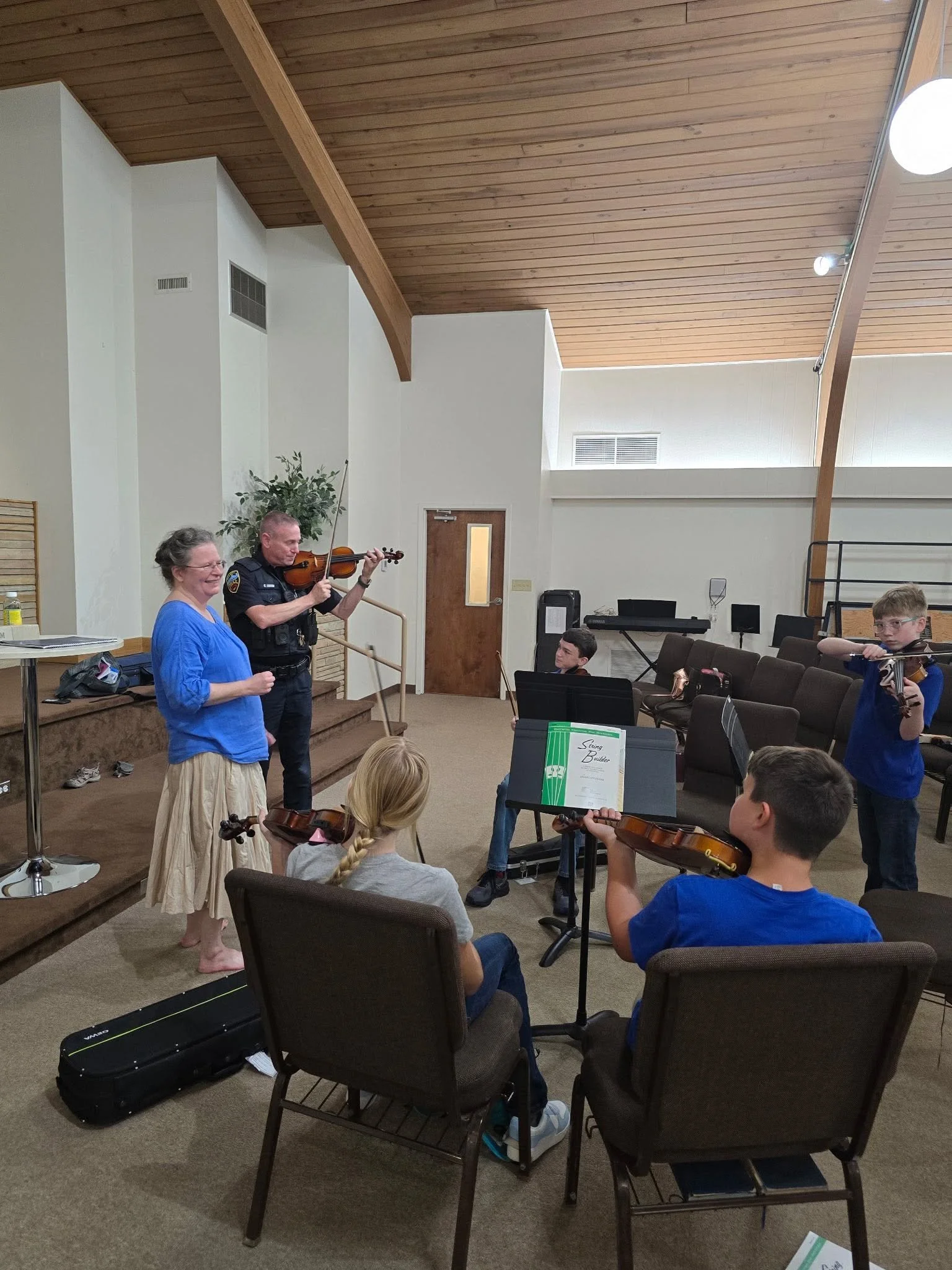 A group of children and two adults in a room with wooden ceiling, practicing or playing string instruments like violins, with music stands and classroom music equipment.