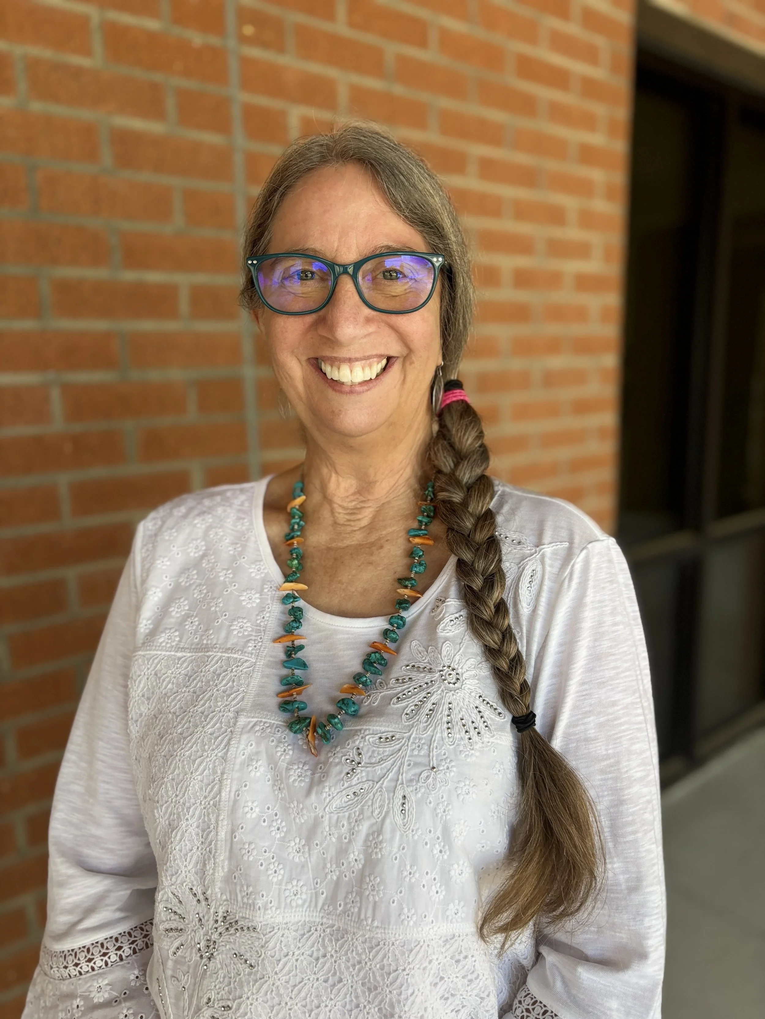 A woman with glasses and a long braid smiling, wearing a white embroidered top and a turquoise and orange beaded necklace, standing in front of a brick wall.