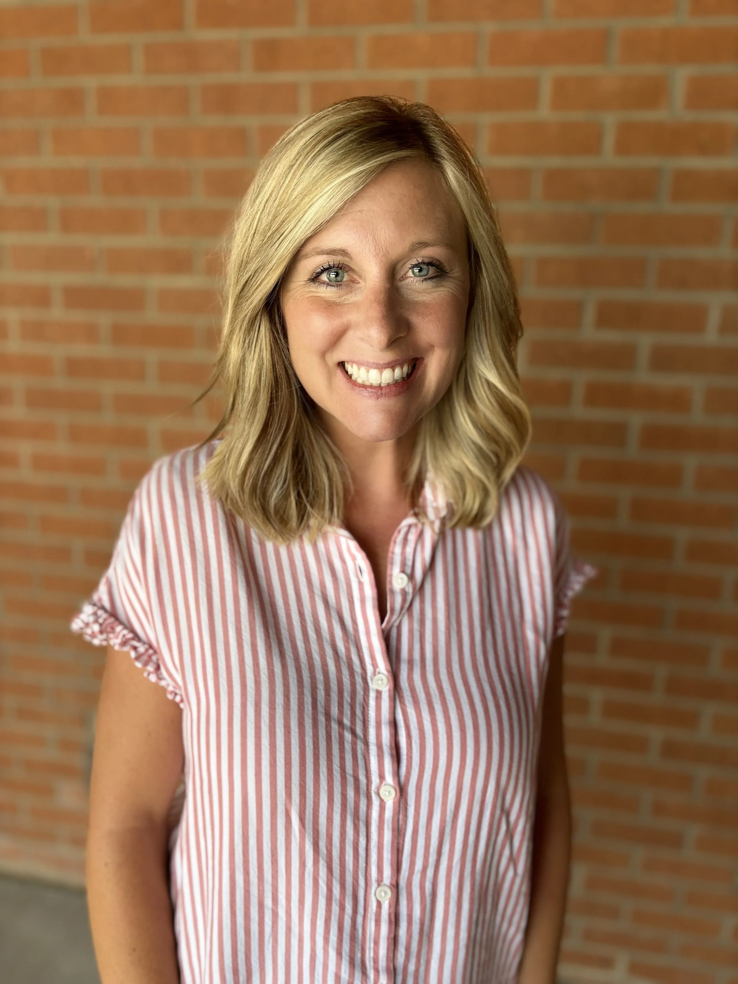 A woman with shoulder-length blonde hair smiling, wearing a red and white striped button-up shirt, standing in front of a brick wall.
