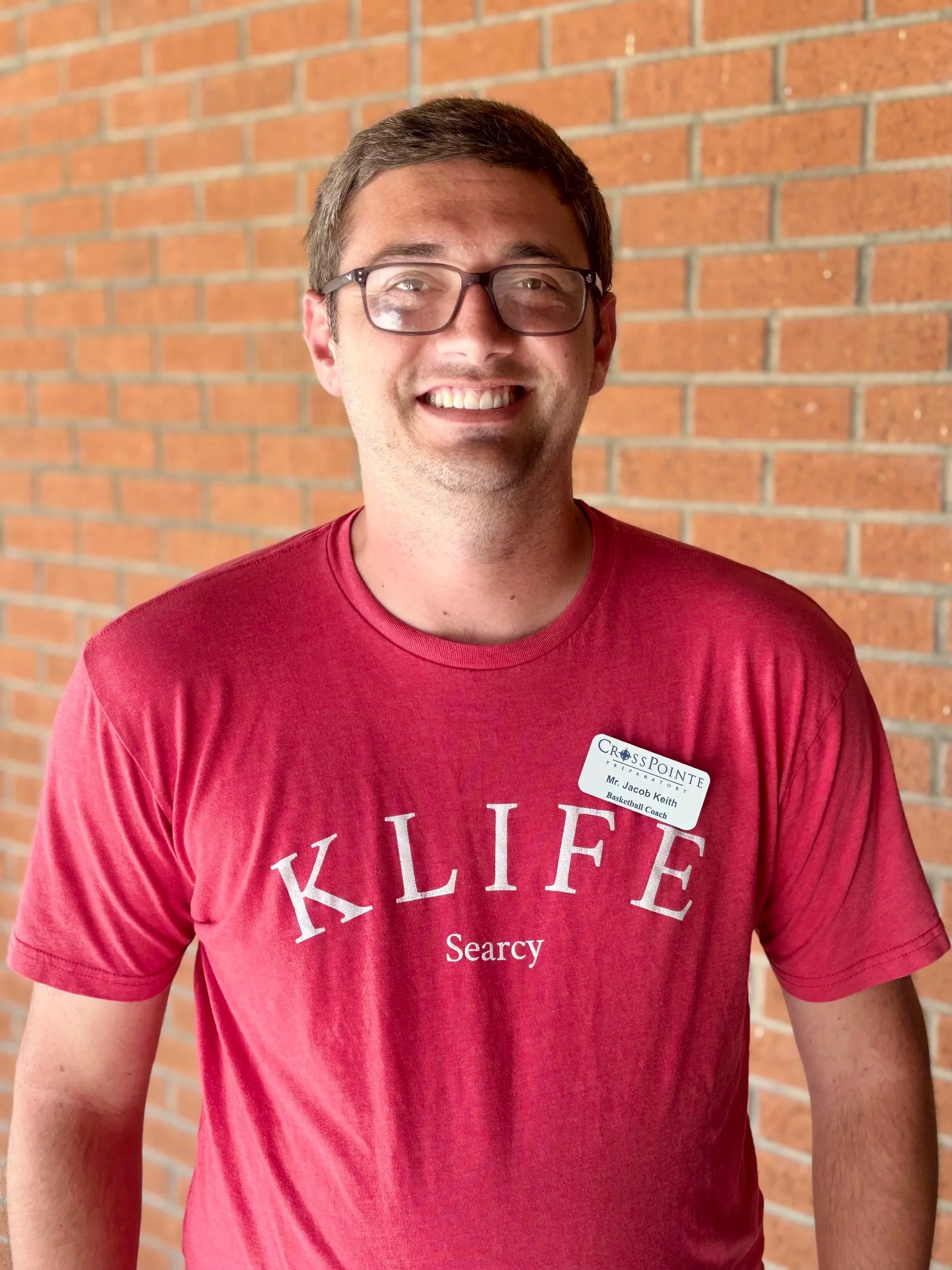 A smiling man wearing glasses and a red T-shirt with white writing, standing against a brick wall. He has a name tag that reads Mr. Jacob Keith, Basketball Coach, from CrossPointe Preparatory.
