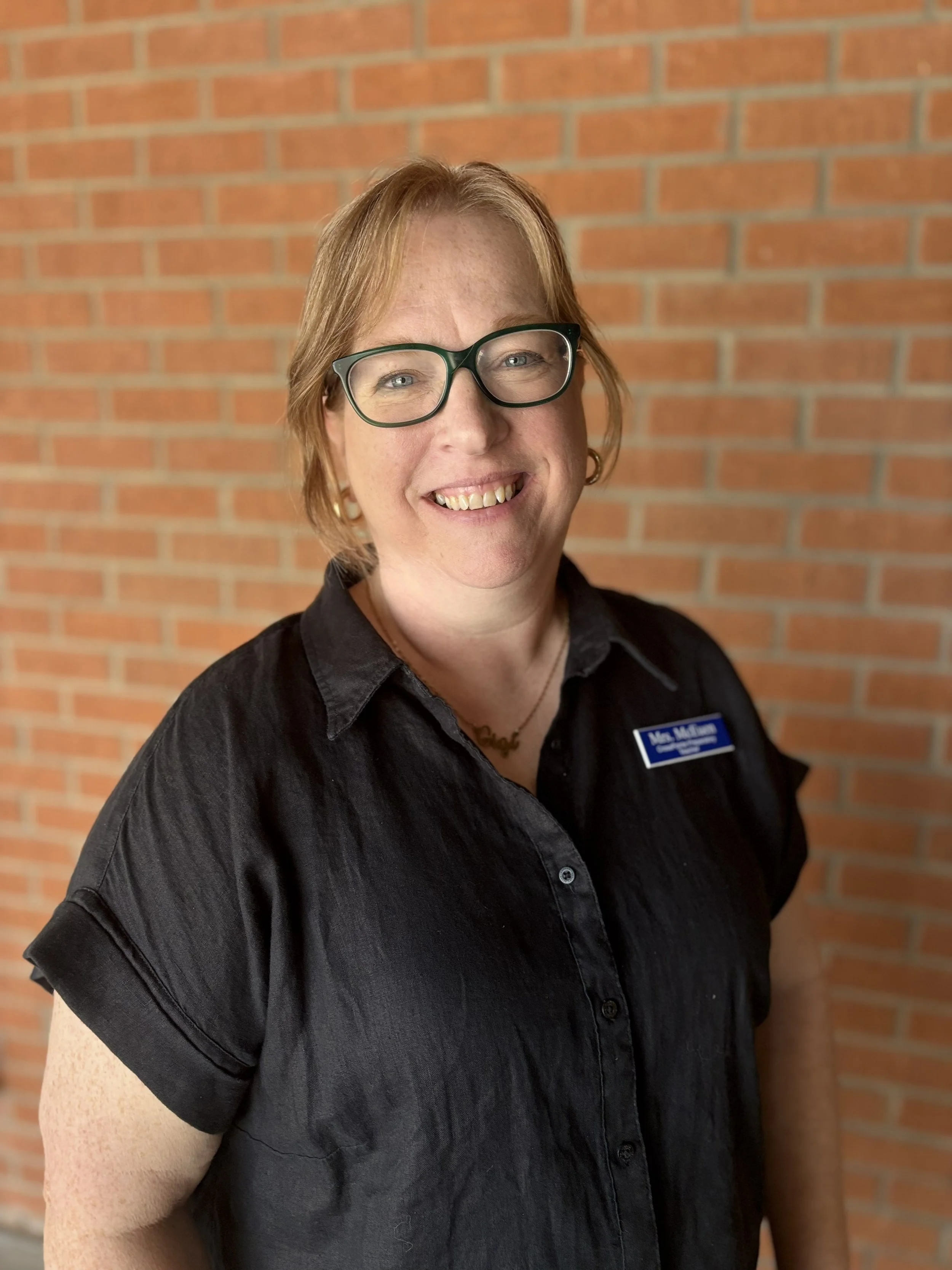 Smiling woman with short reddish hair, glasses, wearing a black shirt with a name tag, standing in front of a brick wall.