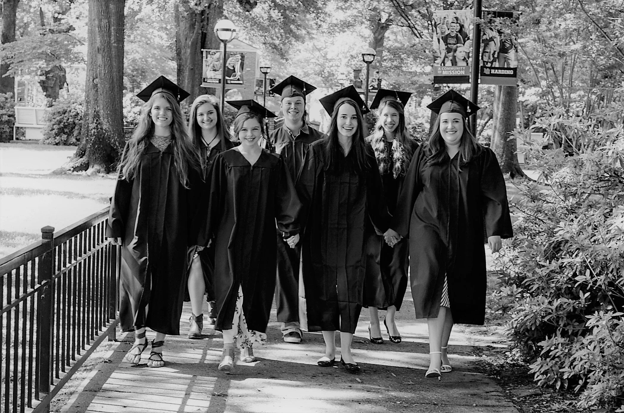 Group of eight young women in graduation caps and gowns walking outdoors on a pathway in a wooded area.
