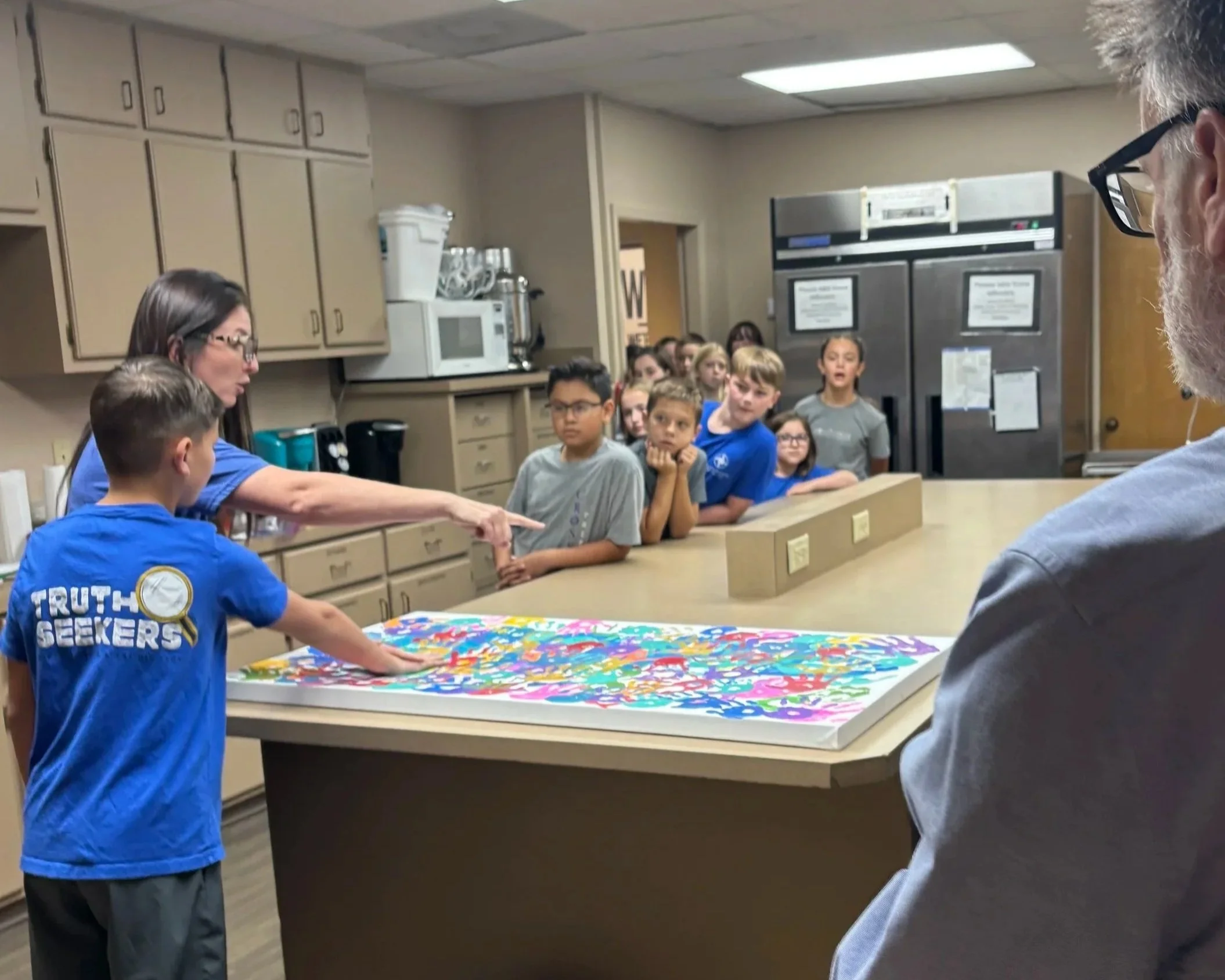 Children gathered around a colorful table in a kitchen or community room, with a woman in blue leading an activity, and a man with glasses observing. There are cabinets, a microwave, and a large refrigerator in the background.