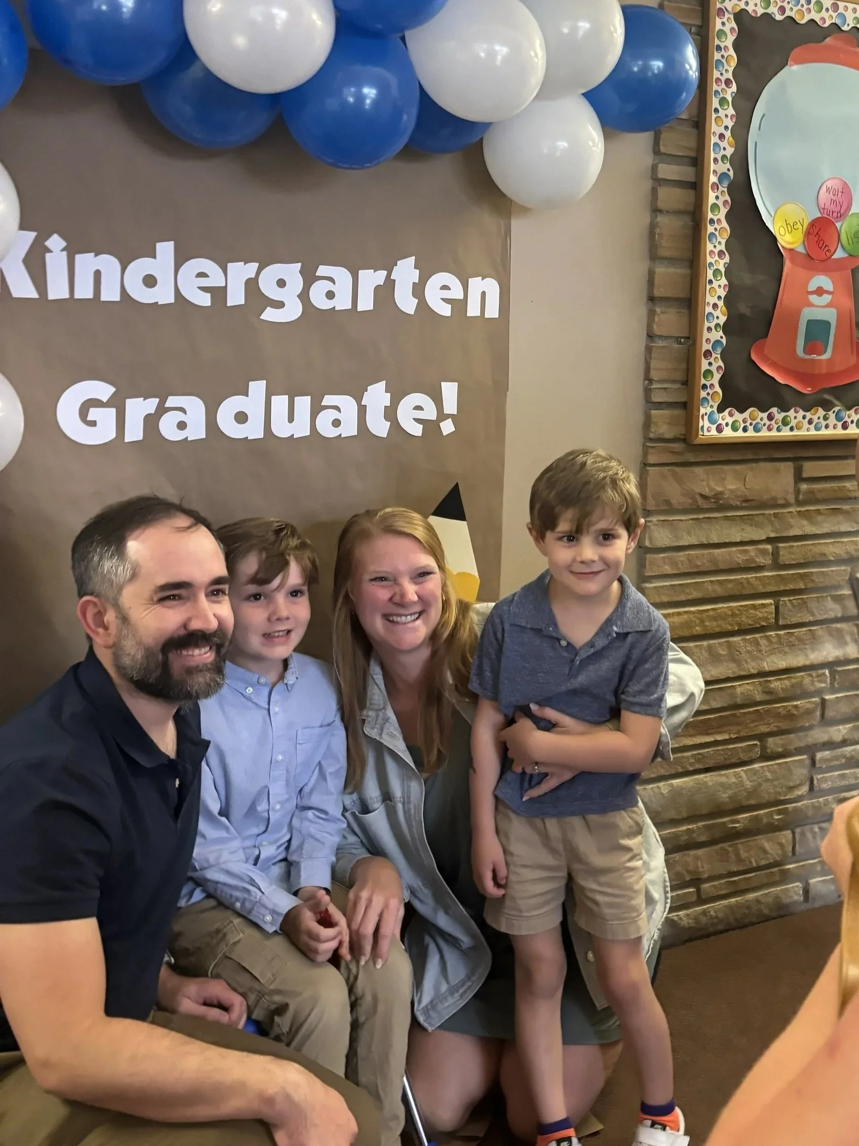 A group of four kids and one adult posing for a photo at a kindergarten graduation event, with a decorative balloon arch and a bulletin board with colorful decorations in the background.