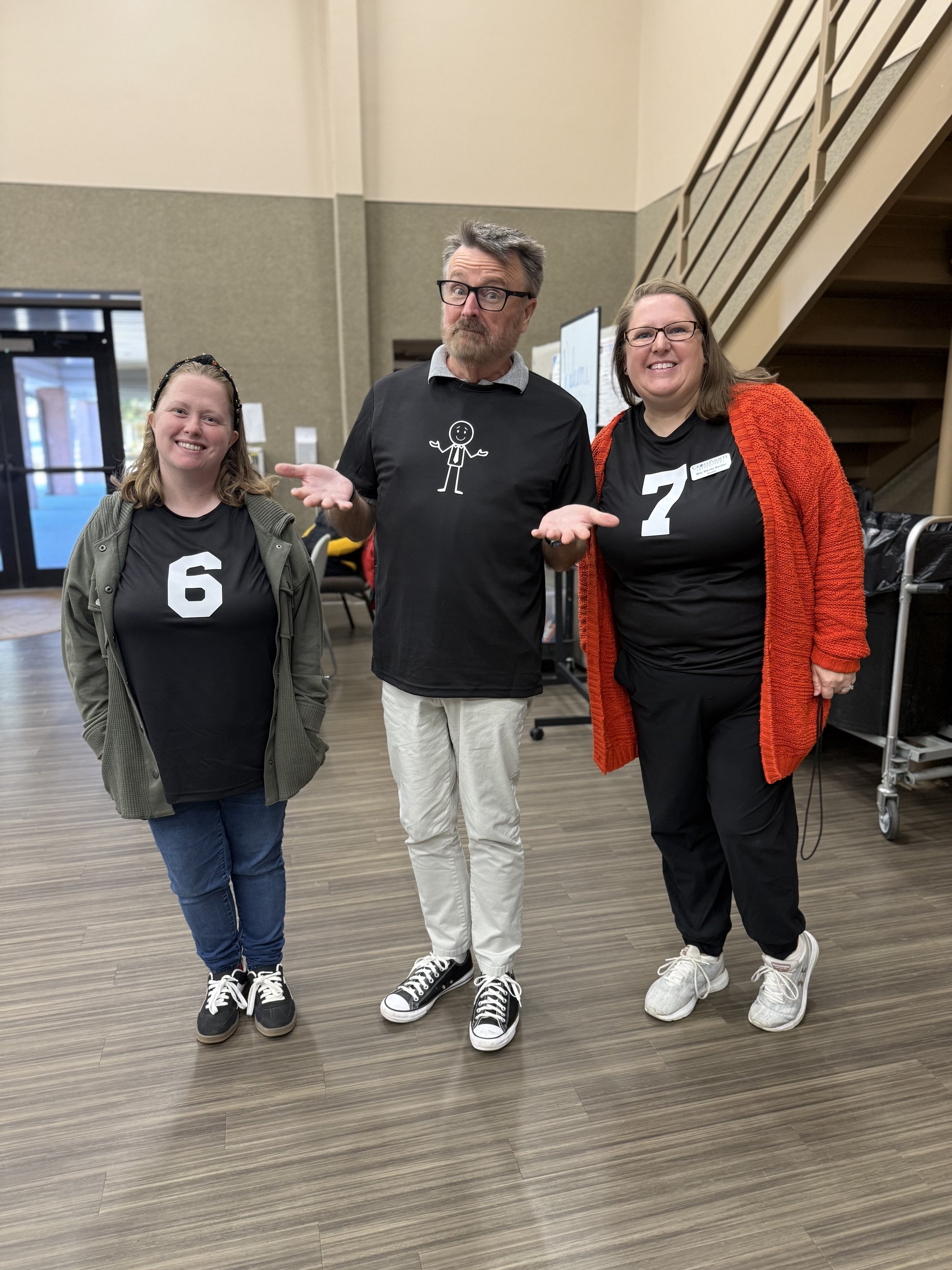 Three people standing inside a building, smiling and posing for the camera. The person in the middle has a puzzled expression, while the other two are smiling. They are wearing black shirts with white numbers and casual clothing.