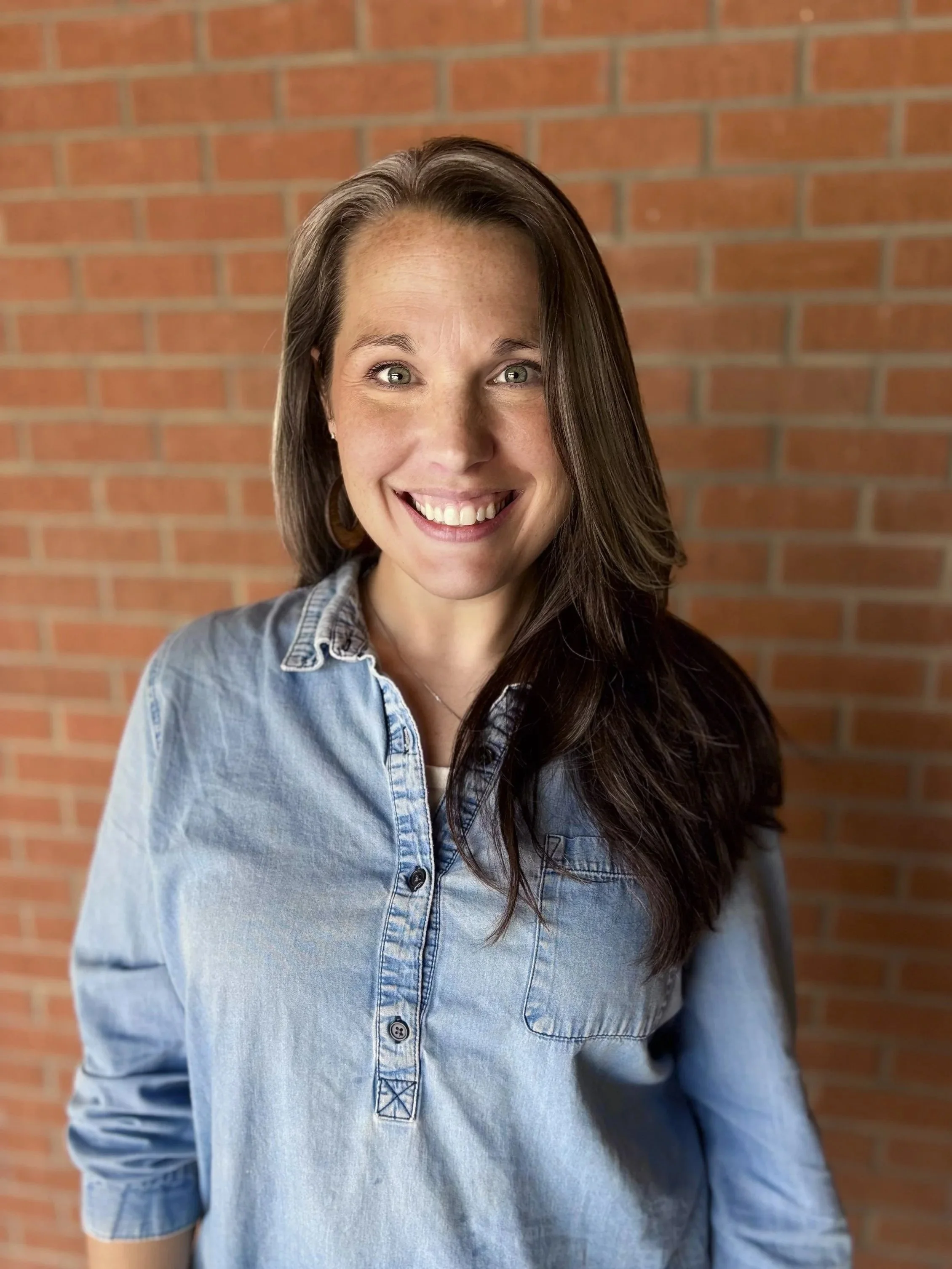 Close-up of a smiling woman with long dark hair, wearing a light blue denim shirt, standing in front of a brick wall.