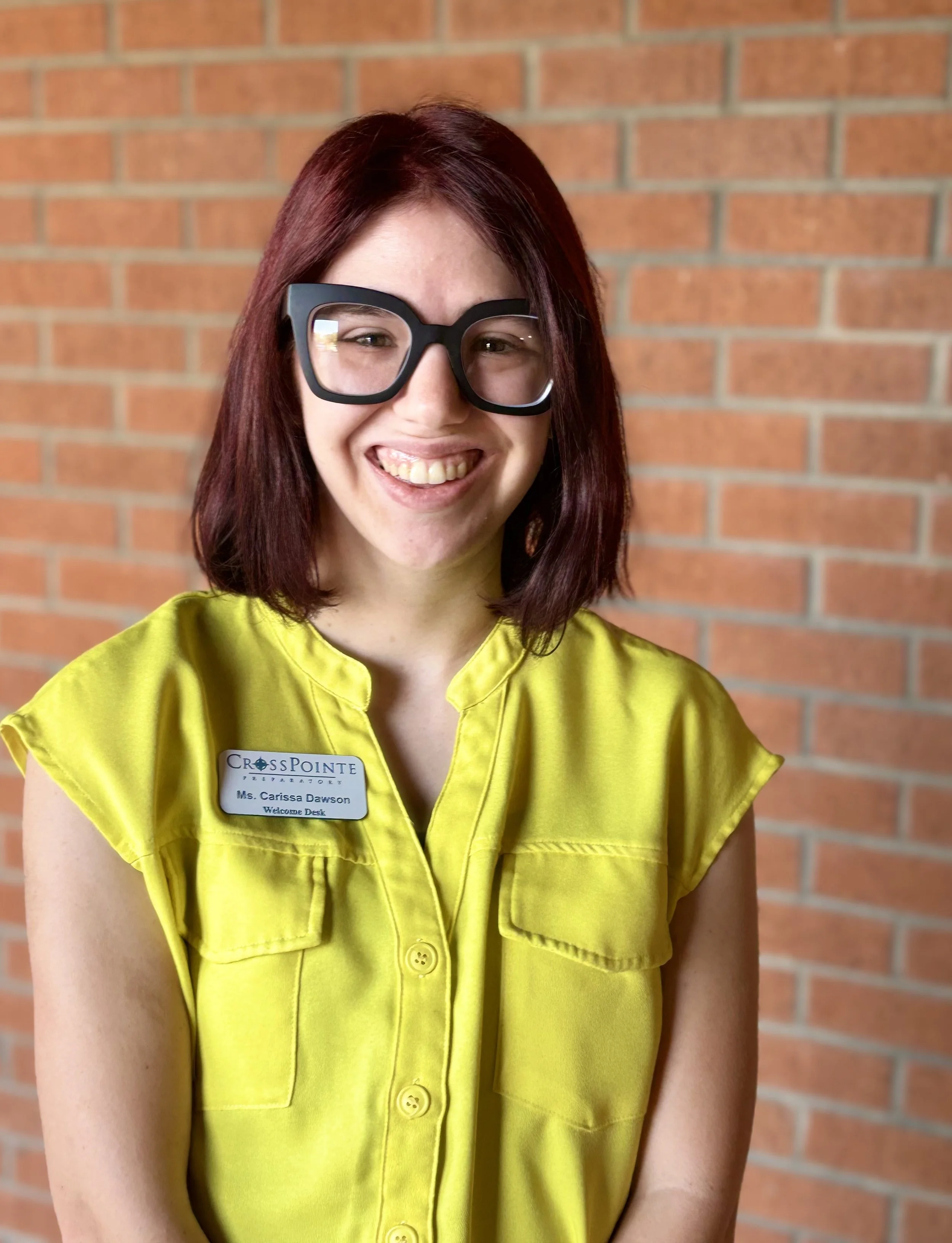 A woman with short reddish-brown hair wearing black glasses and a bright yellow shirt, smiling, standing in front of a brick wall. She has a name tag that reads "Ms. Carissa Dawson, Welcome Desk," from CrossPointe Megastore.