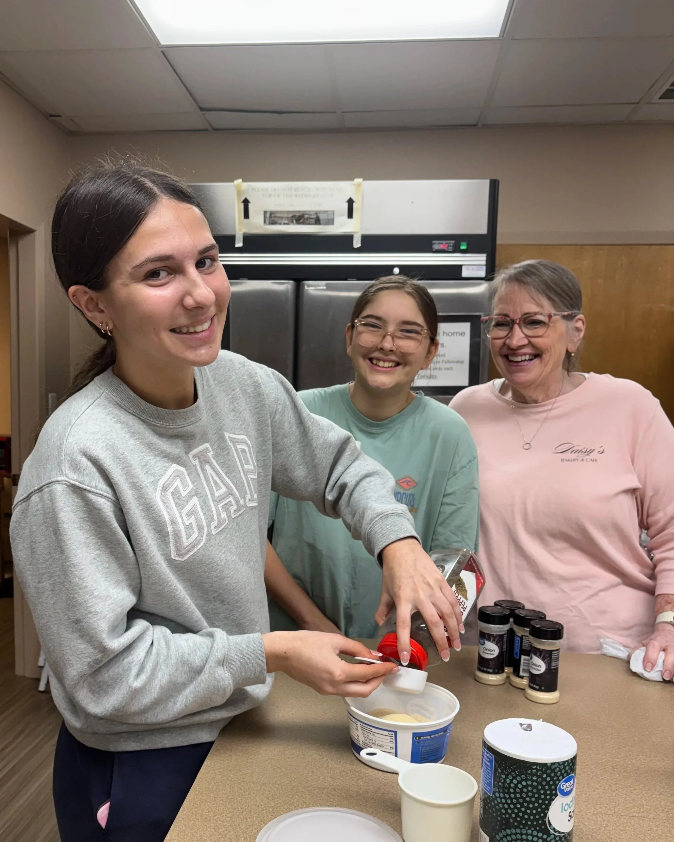Three women preparing ice cream at a counter with tubs of ice cream and toppings, in an indoor setting.