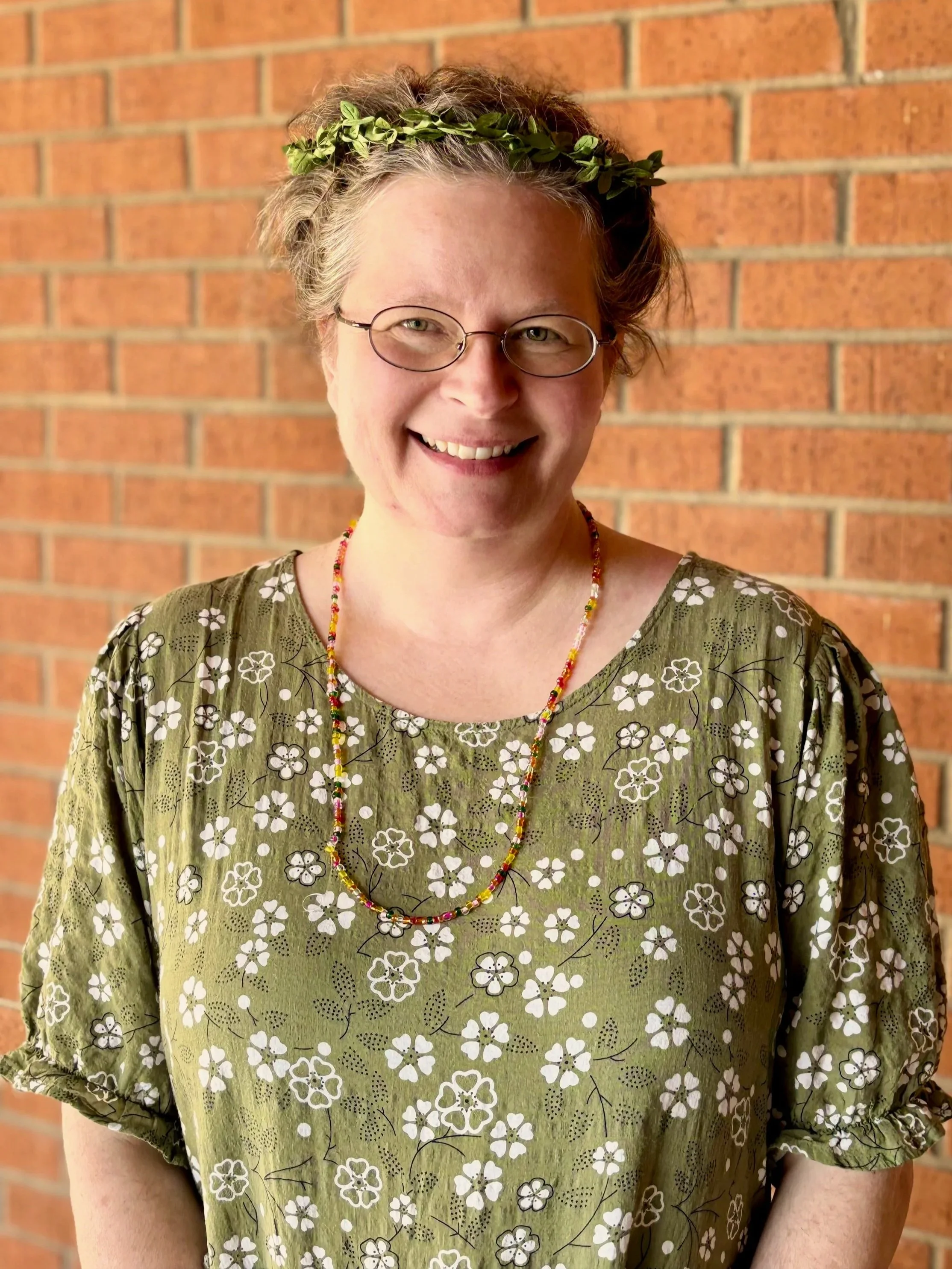 A woman with short blonde hair, wearing glasses, a green floral blouse, a colorful beaded necklace, and a green leafy headband, standing in front of a brick wall.