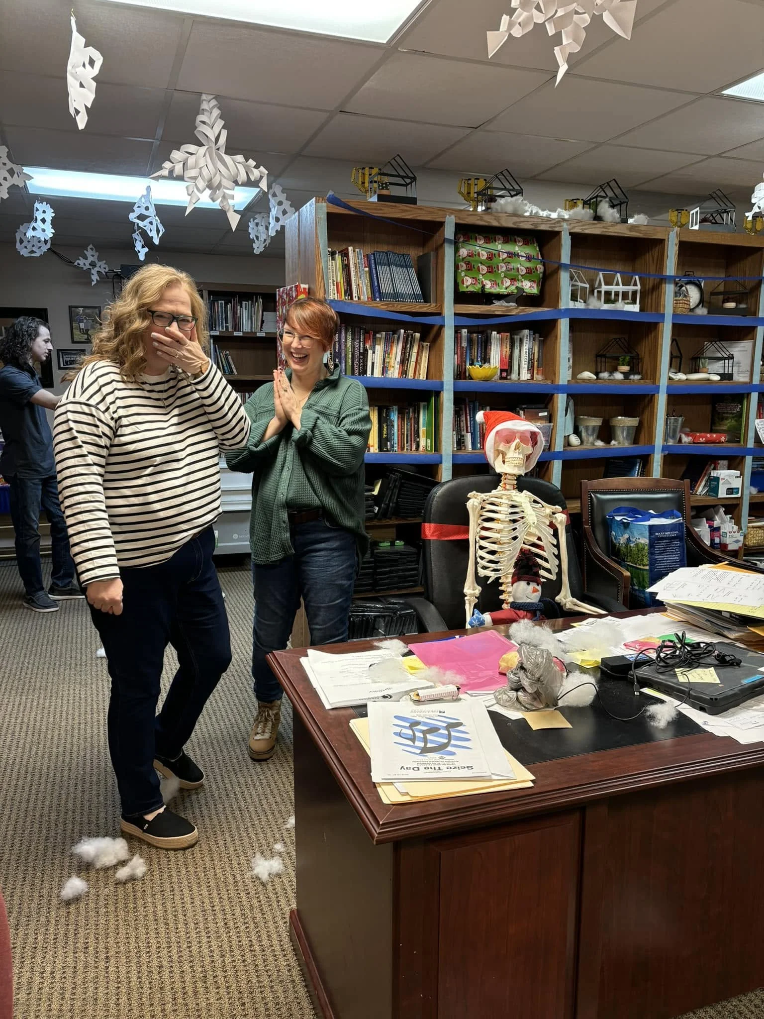Two women standing in an office decorated with paper snowflakes and paper houses hanging from the ceiling, with a skeleton dressed as Santa Claus on the desk.
