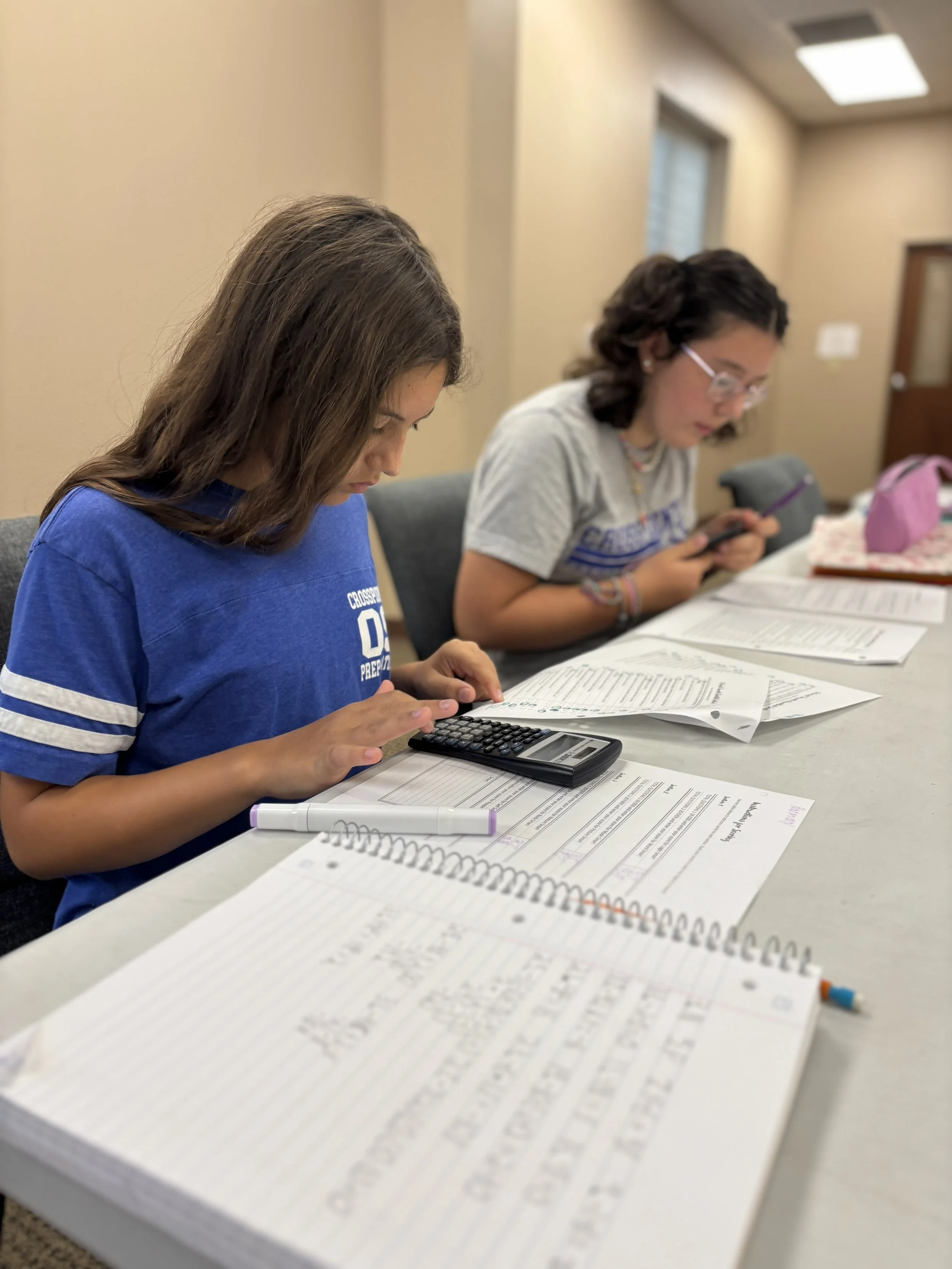 Two teenage girls studying with papers, a calculator, and a notebook on a long table in a classroom or library setting.