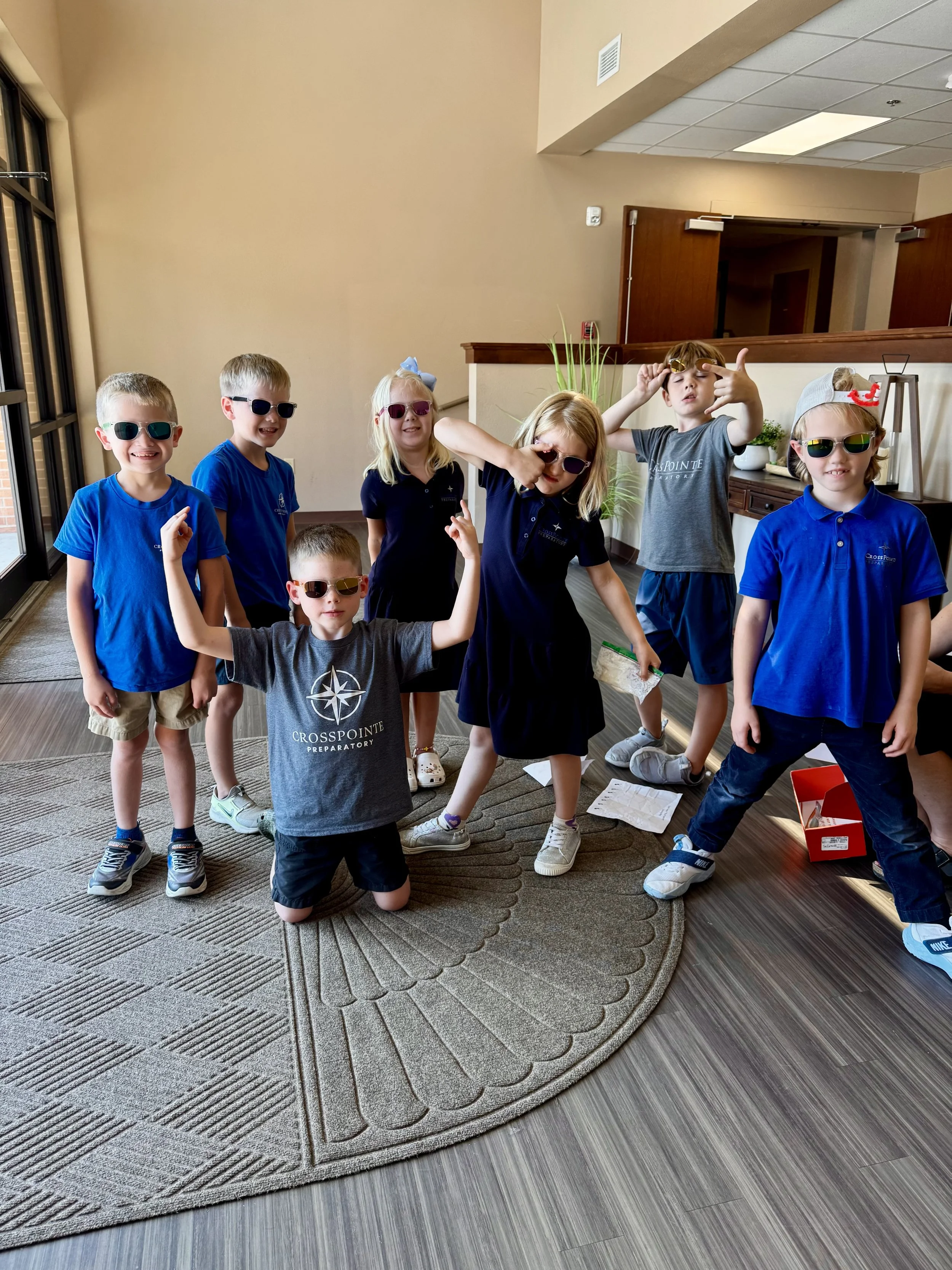 Seven children wearing sunglasses and casual clothing, standing in a room with beige walls and a patterned carpet. Some children are making gestures and posing playfully, while others are smiling.