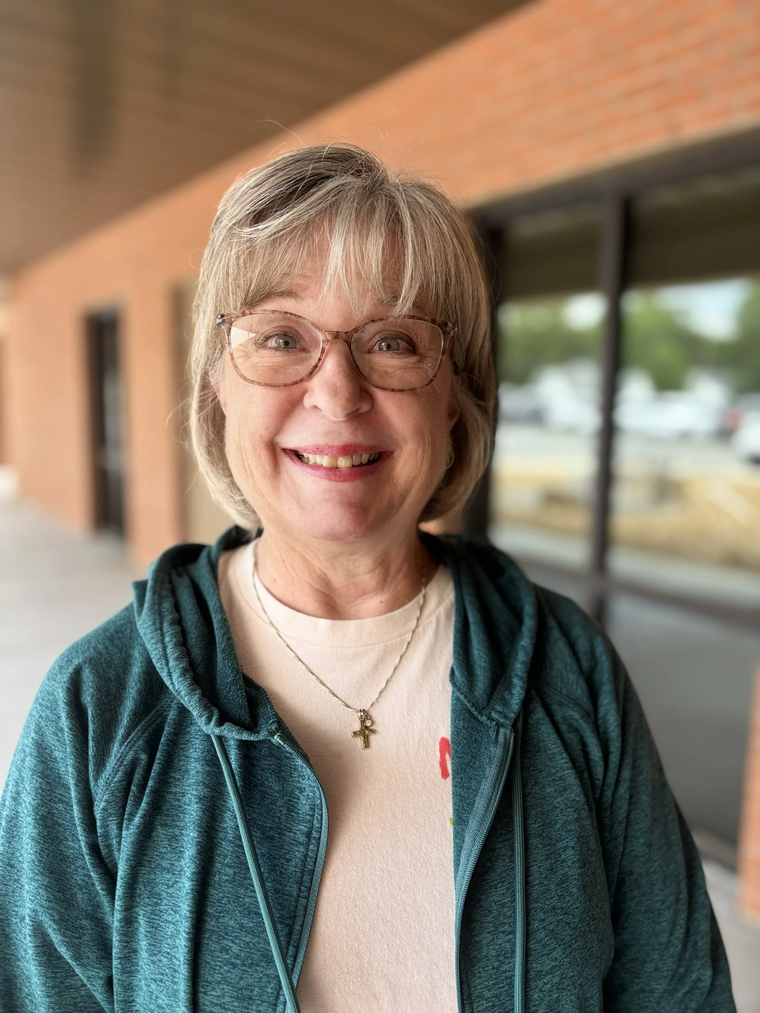 Close-up of a smiling woman with gray hair, wearing glasses, a white t-shirt, a teal zip-up hoodie, and a gold cross necklace, standing indoors near large windows with a brick wall in the background.
