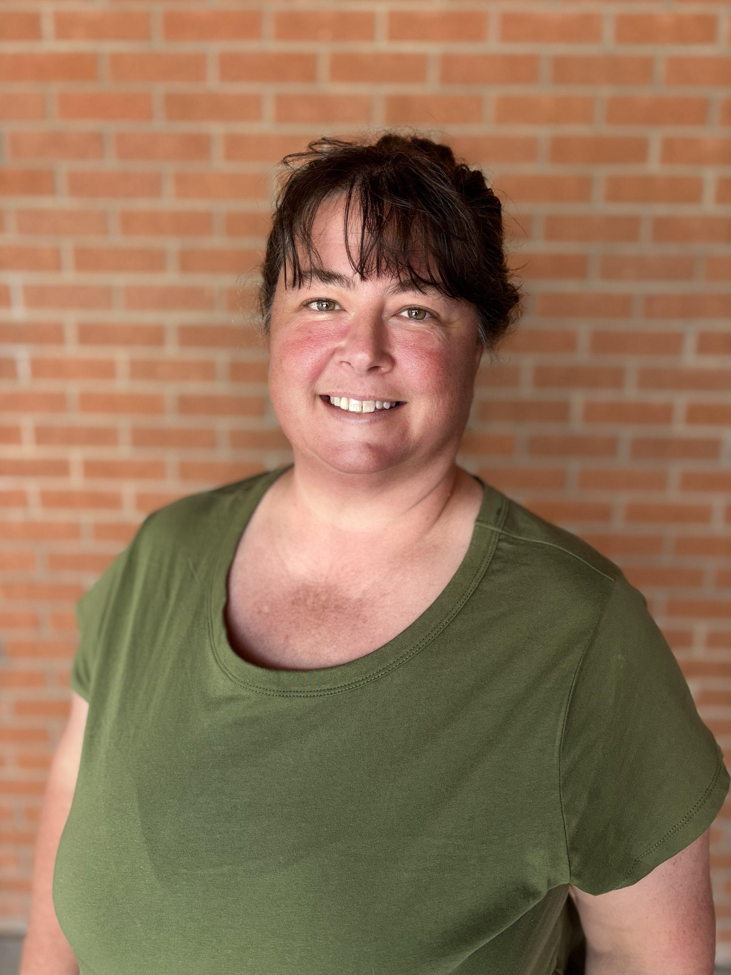 Close-up of a smiling woman wearing a green shirt with a brick wall background.