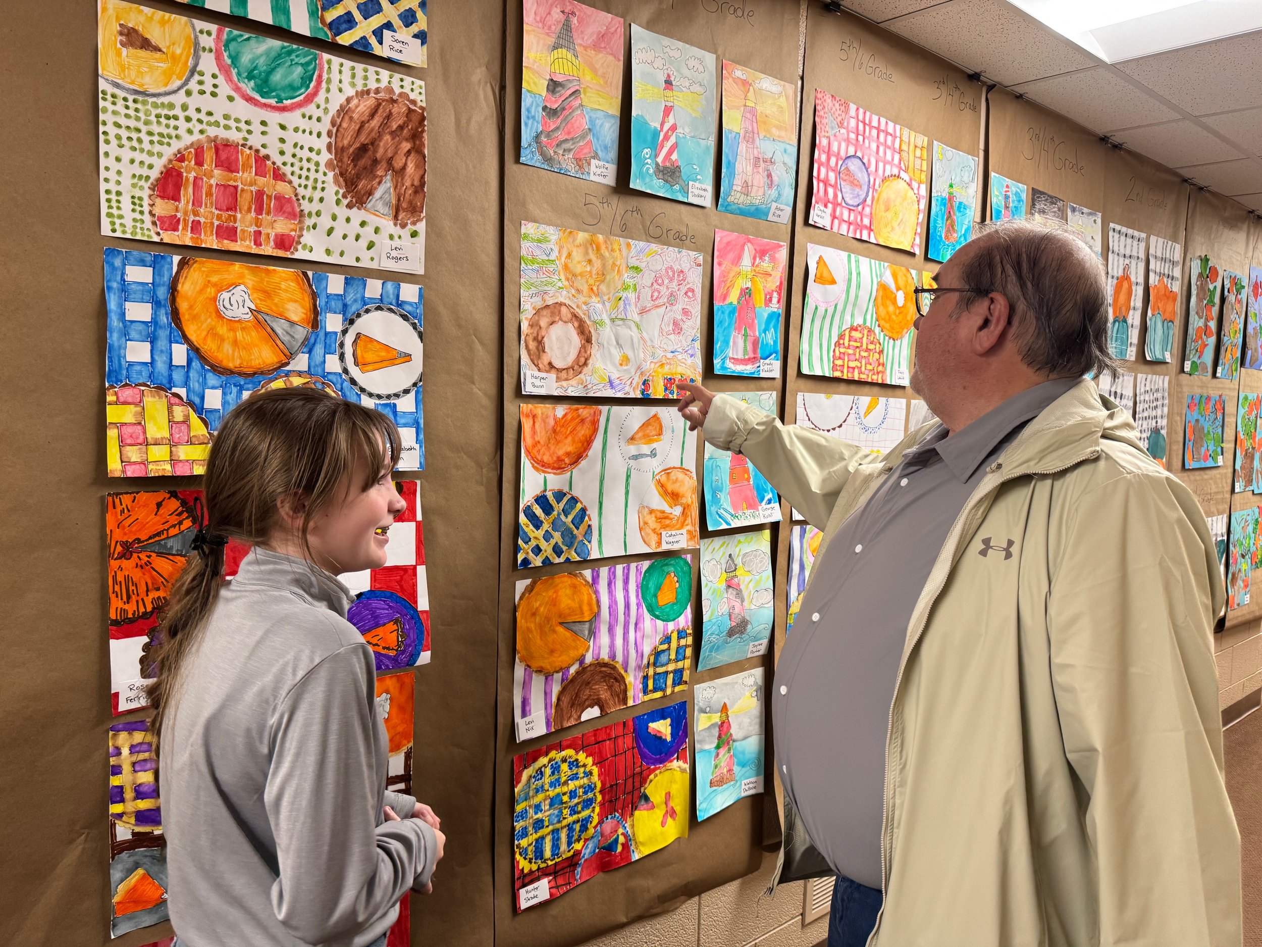 A man and a girl are looking at colorful children's artwork displayed on a wall in an educational setting. The artwork includes paintings of pumpkins, lighthouses, and abstract designs, arranged in rows with labels indicating different grade levels.