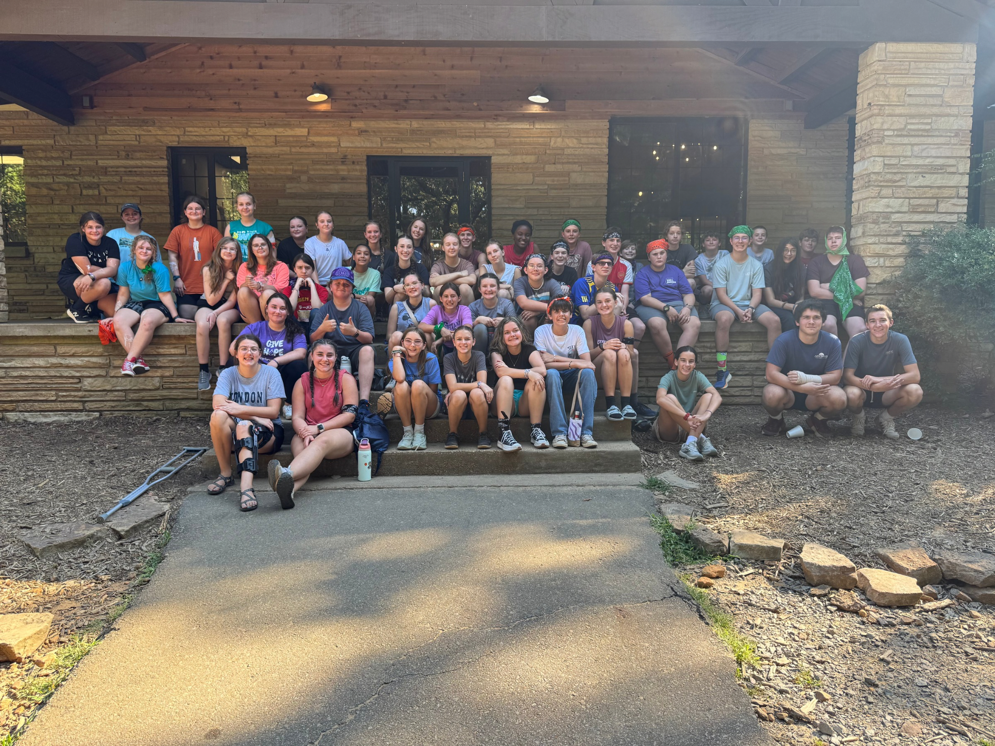 Group of children and teenagers sitting and standing outside in front of a building with wood and brick exterior, some with bandanas, smiling for the photo.