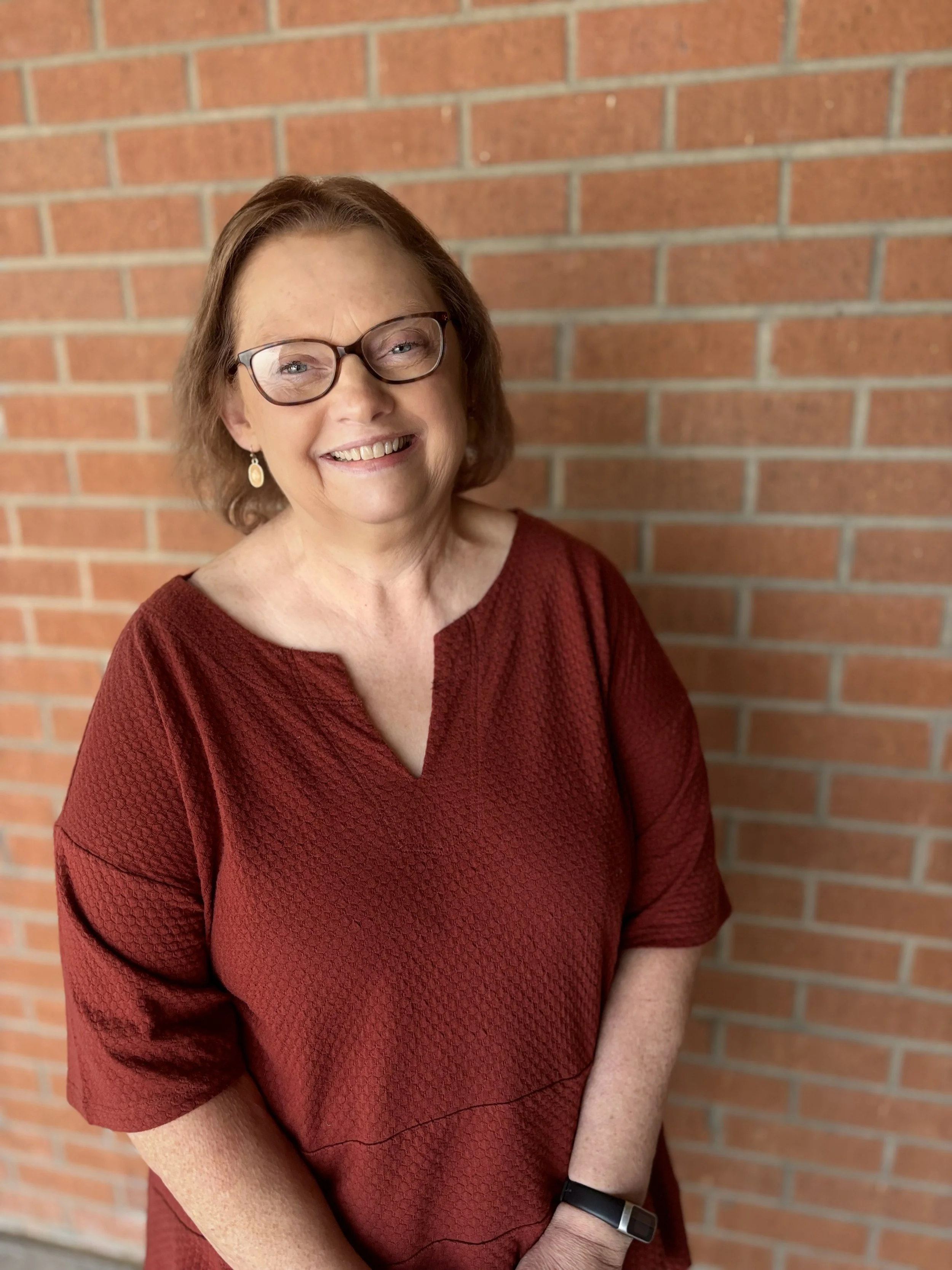 A smiling middle-aged woman with glasses, wearing a red textured top, standing in front of a brick wall.