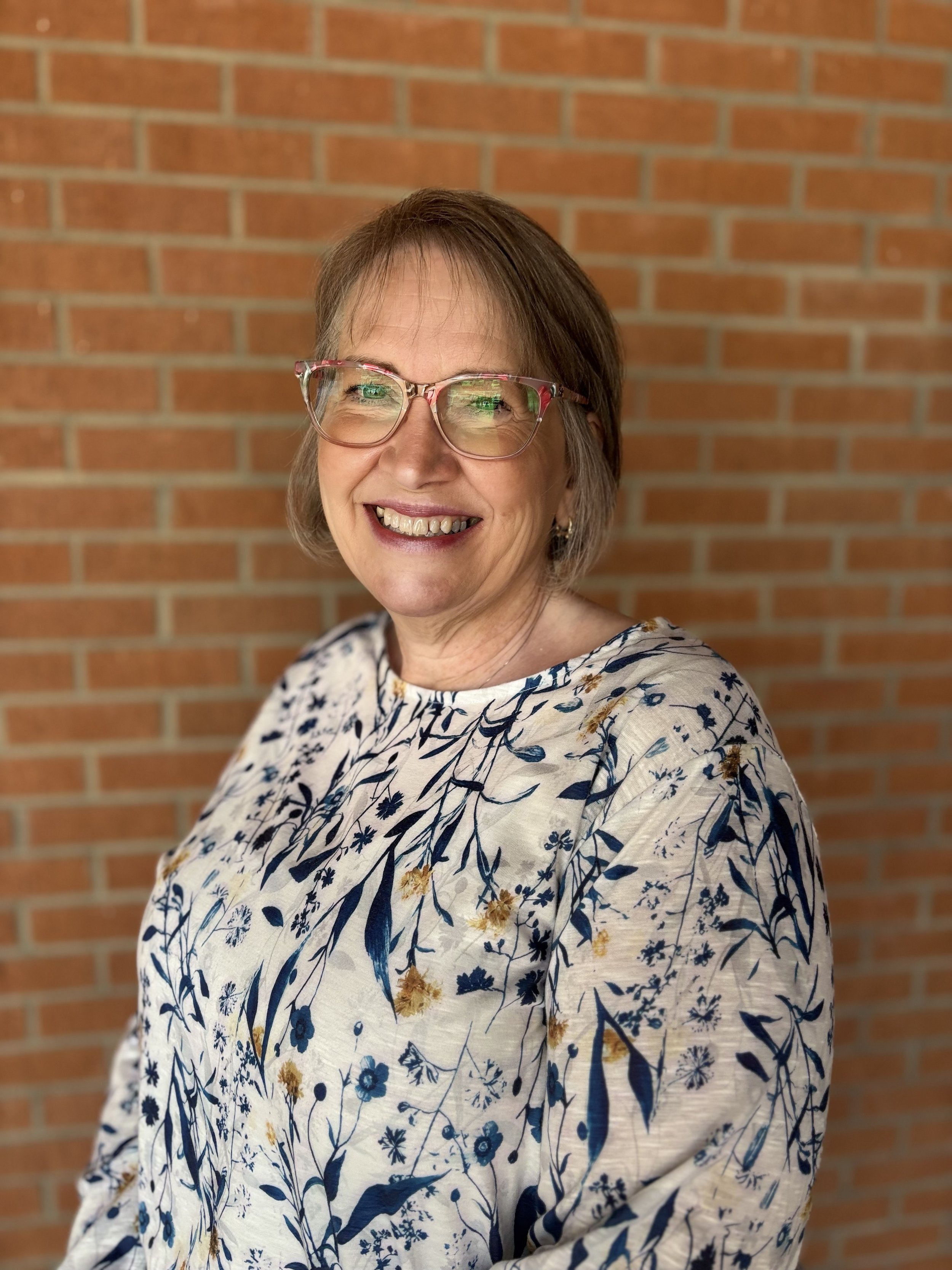 A middle-aged woman with short brown hair and glasses, smiling, wearing a floral blouse, standing against a brick wall background.