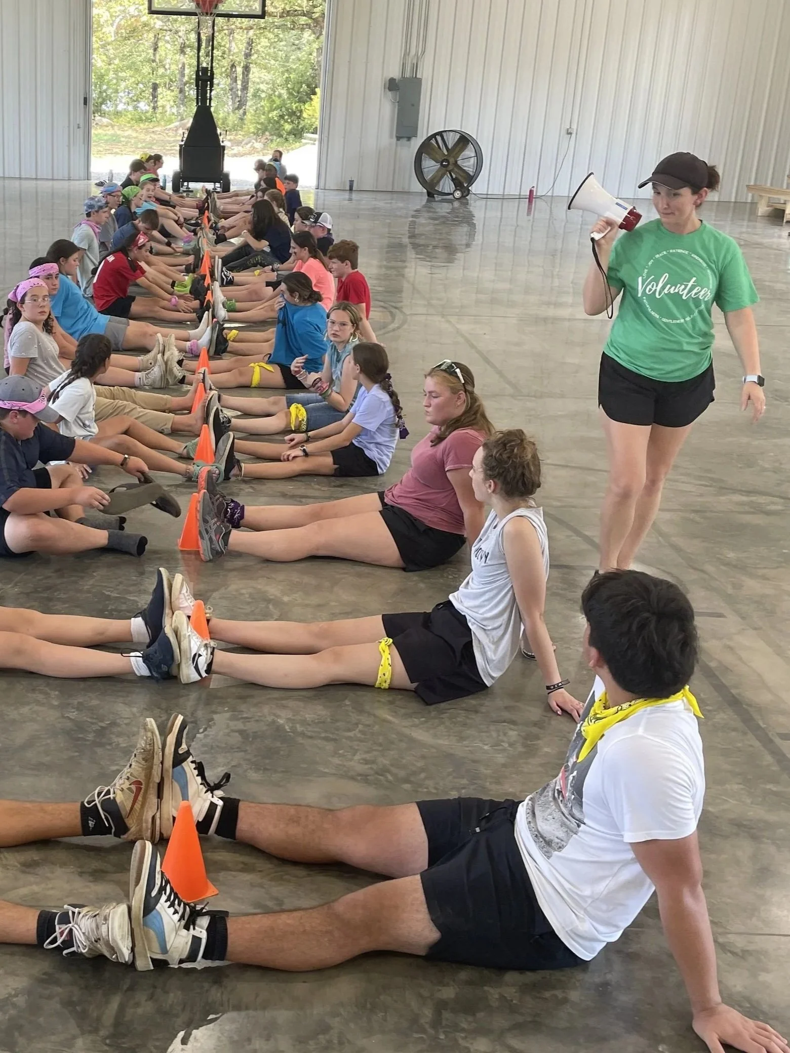 A group of children sitting on the floor in a gymnasium, stretching with their legs extended and hands reaching forward, supervised by a volunteer with a megaphone.