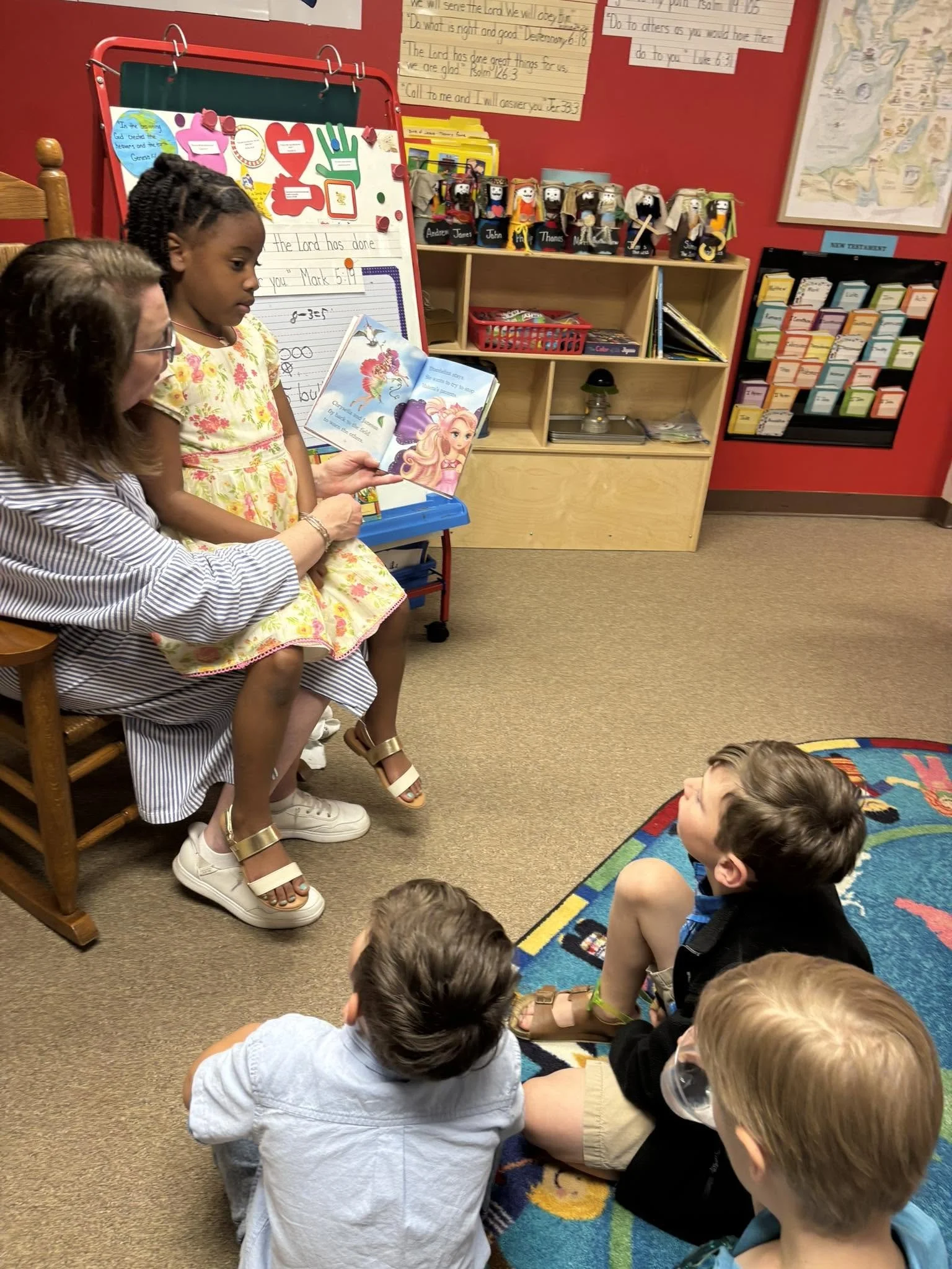 A classroom scene with a teacher and young students sitting on the floor and on chairs. The teacher is showing a children's book to a girl in a yellow floral dress. Several children are attentively watching, sitting cross-legged on a colorful rug. Background includes a red wall with educational posters, books, and decorative objects.