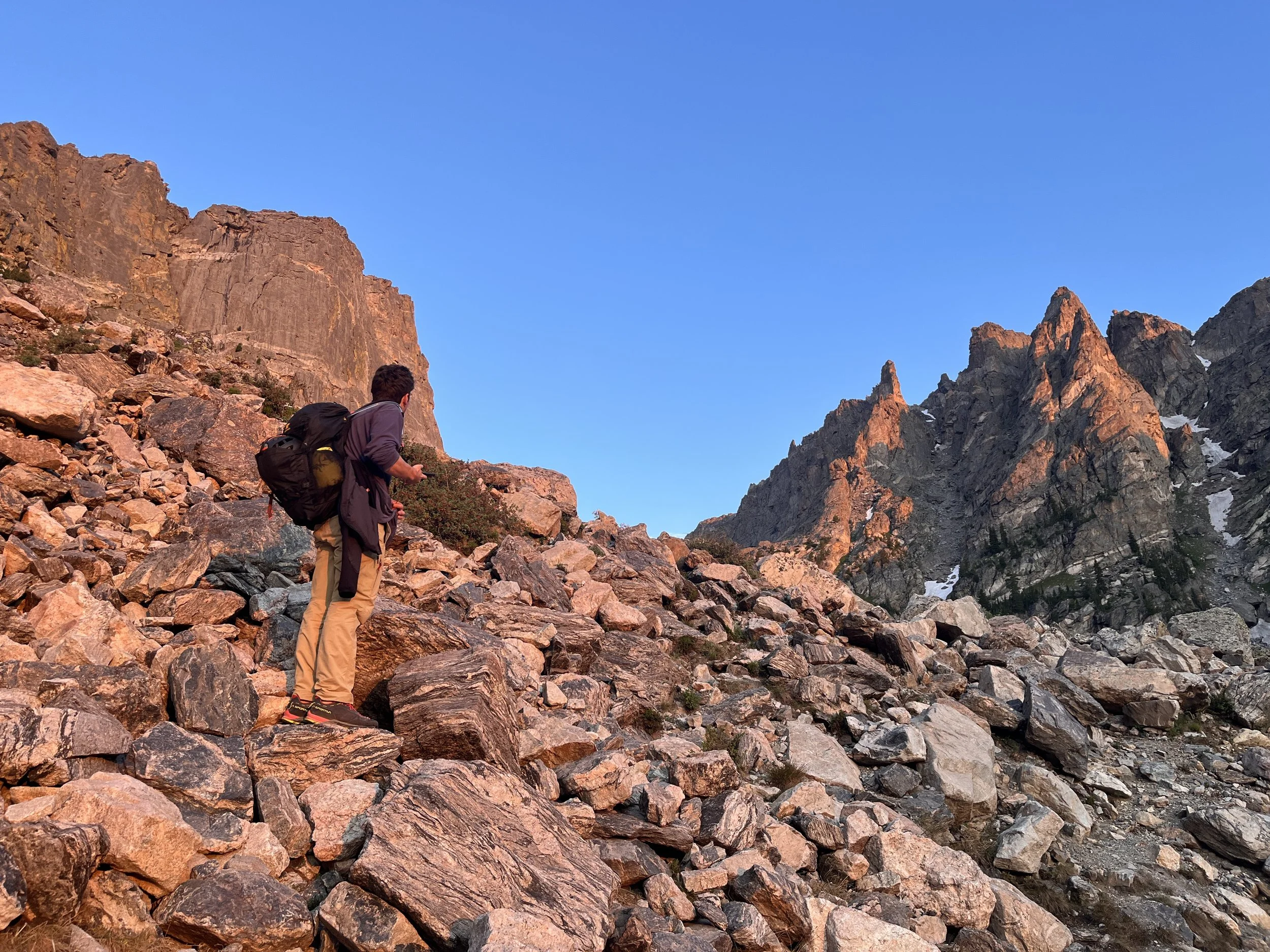 A person with a backpack standing on rocky terrain in a mountainous area under a clear blue sky.