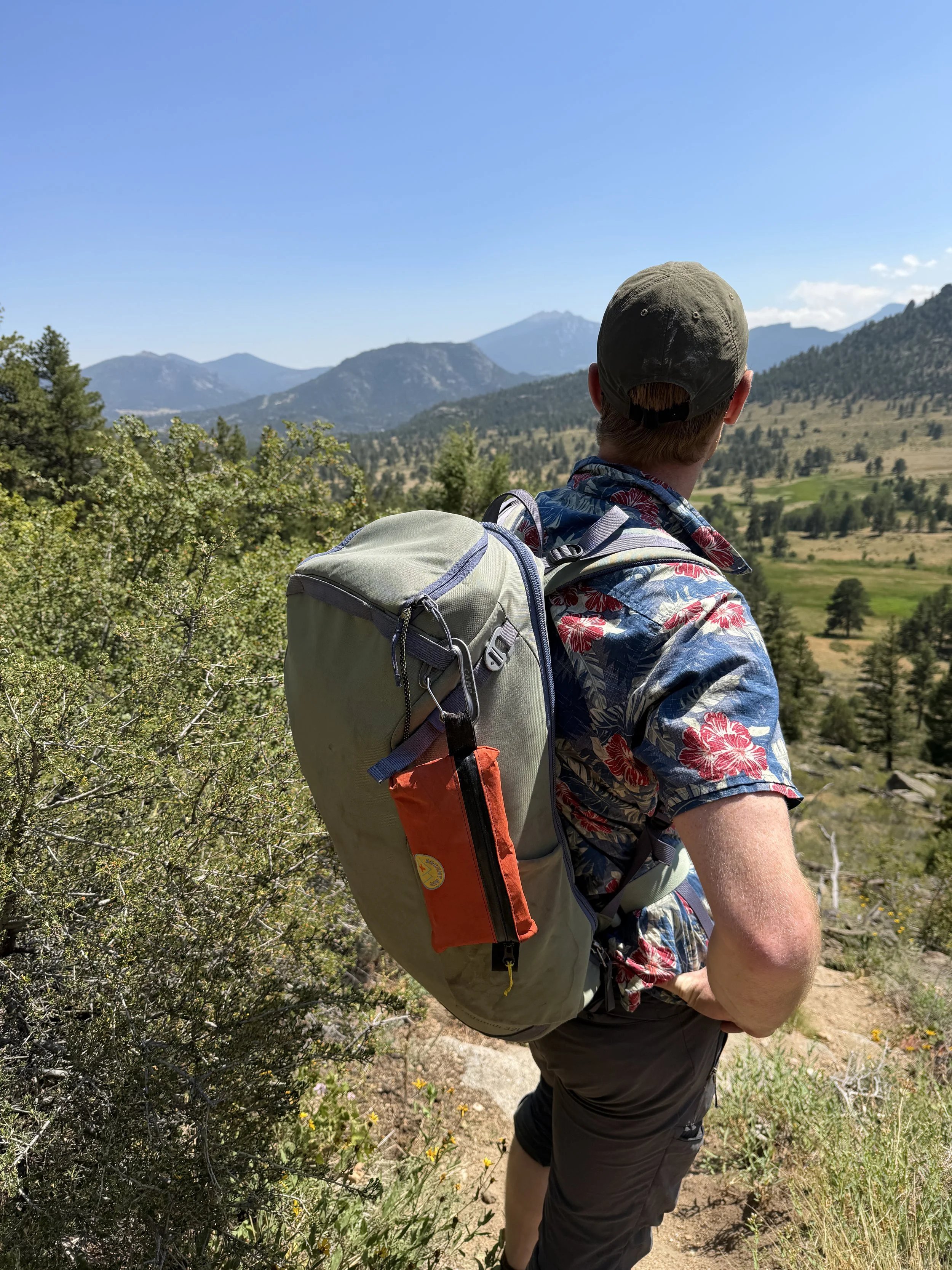 A man hiking on a trail in a mountainous landscape with trees and shrubs, wearing a floral shirt, hat, and backpack.