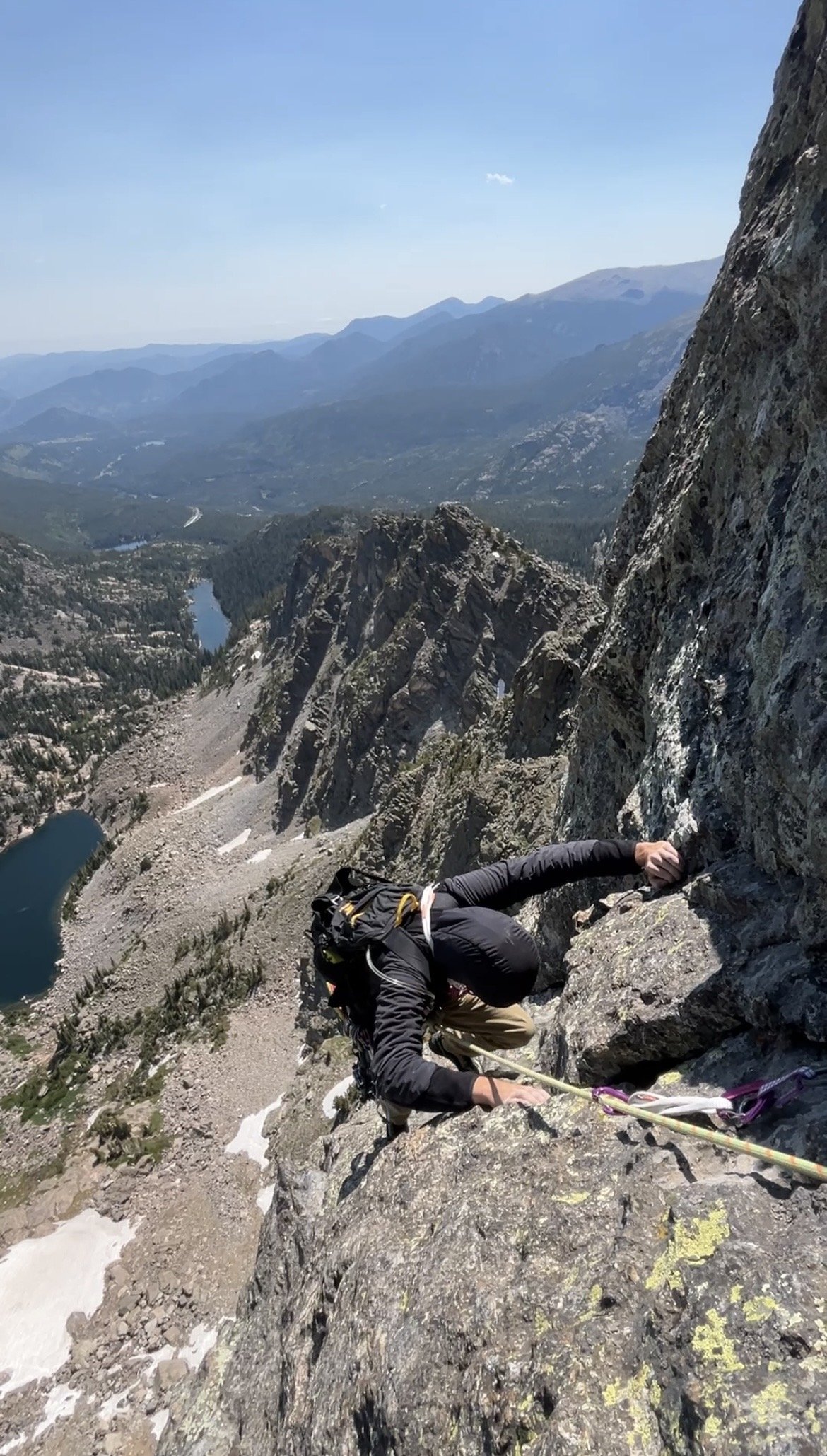 A person rock climbing on a steep mountain face with a scenic mountain valley and lake below.