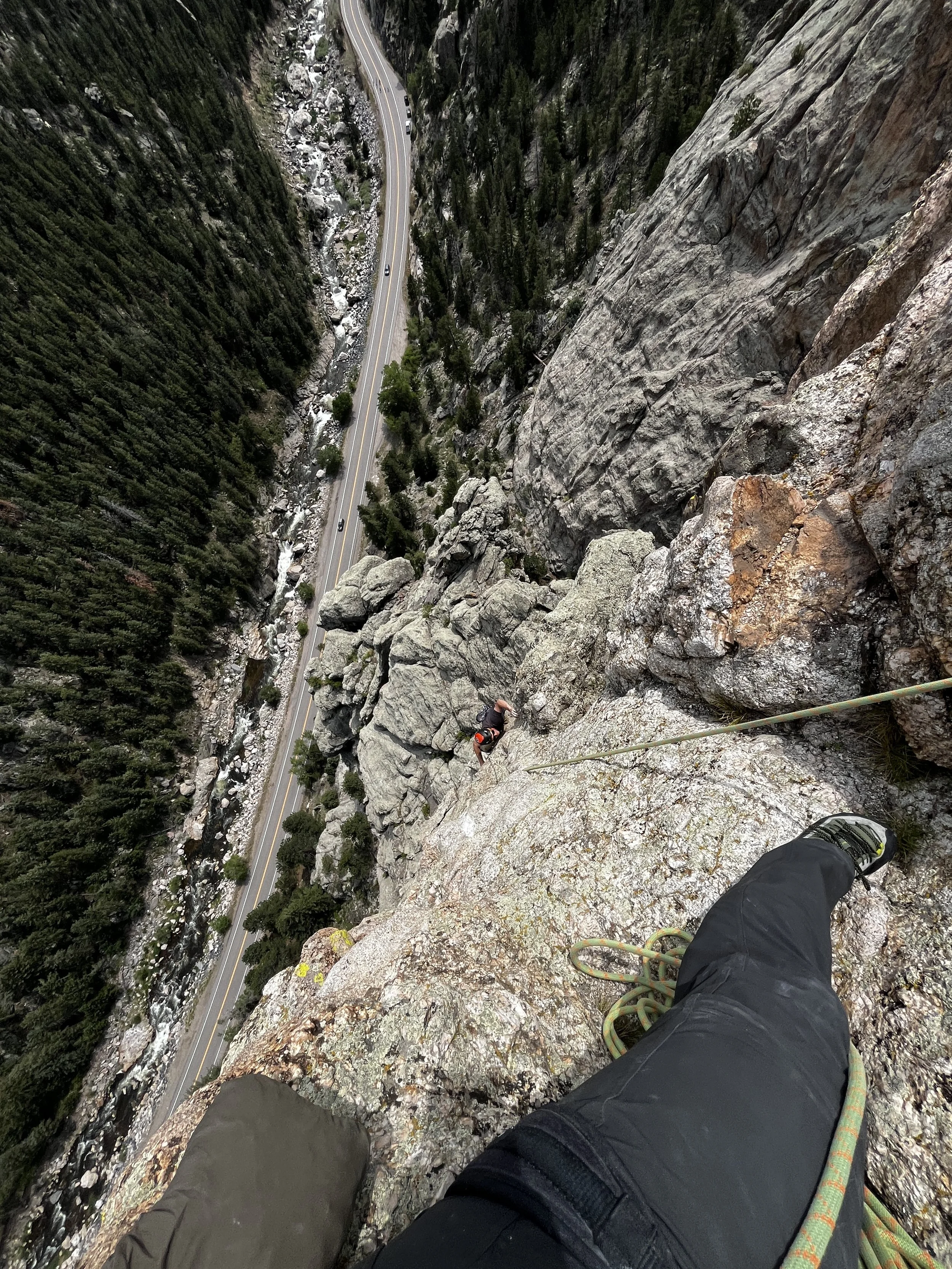 Climber ascending a steep rock face on a mountain, with a valley and a winding road below, taken from the climber's perspective.